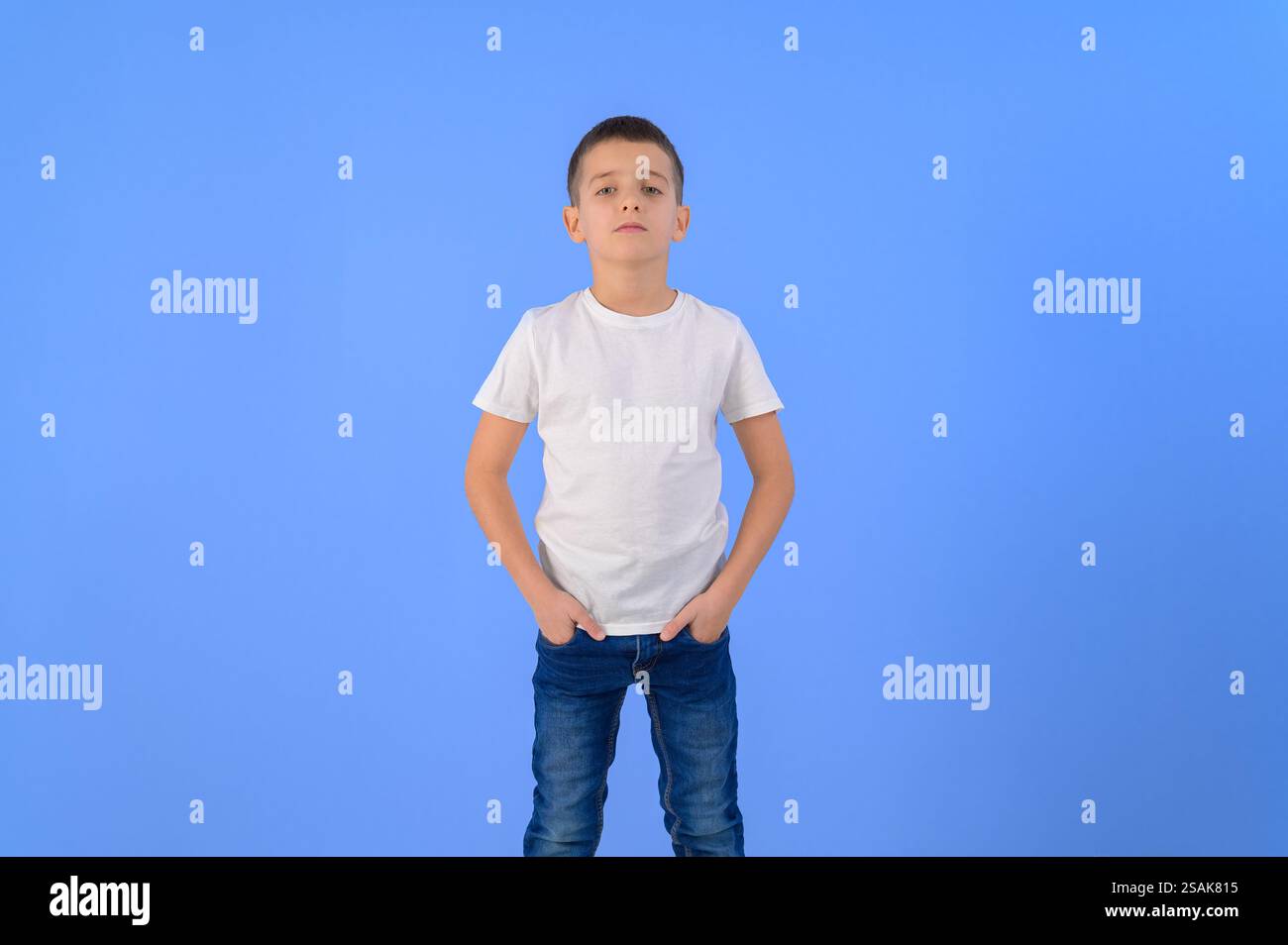 Ritratto di un ragazzo serio e simpatico con le mani in tasca in piedi con sicurezza su sfondo blu isolato Foto Stock