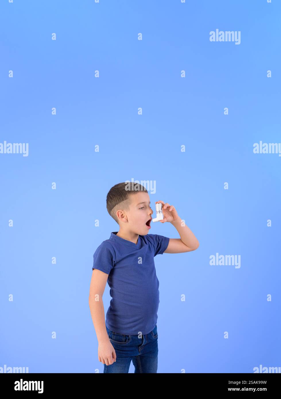 Ritratto di un bambino di sesso maschile che inala attraverso la pompa per asma mentre è in piedi su uno sfondo blu isolato Foto Stock