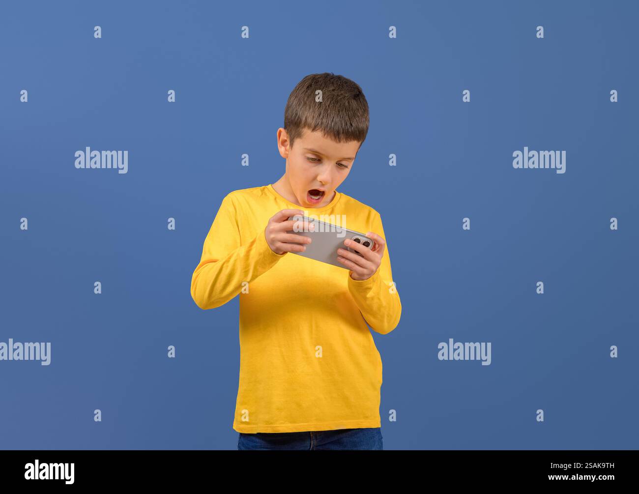 Ritratto di un ragazzo carino con la bocca aperta che gioca a videogame su un cellulare su sfondo blu Foto Stock