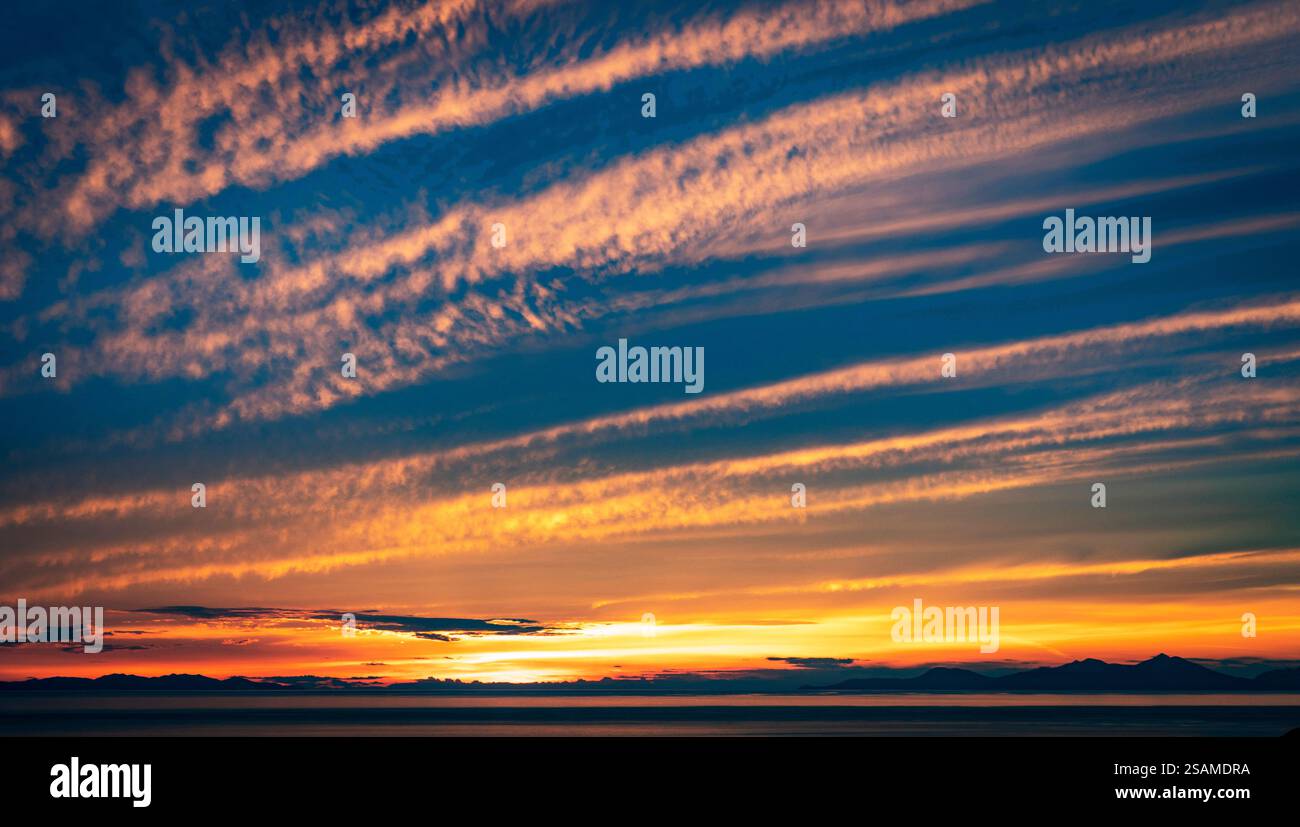 Uno splendido tramonto su un mare calmo, caratterizzato da vivaci sfumature arancioni e blu nel cielo con nuvole di neve. L'orizzonte è fiancheggiato da montagne lontane, c Foto Stock