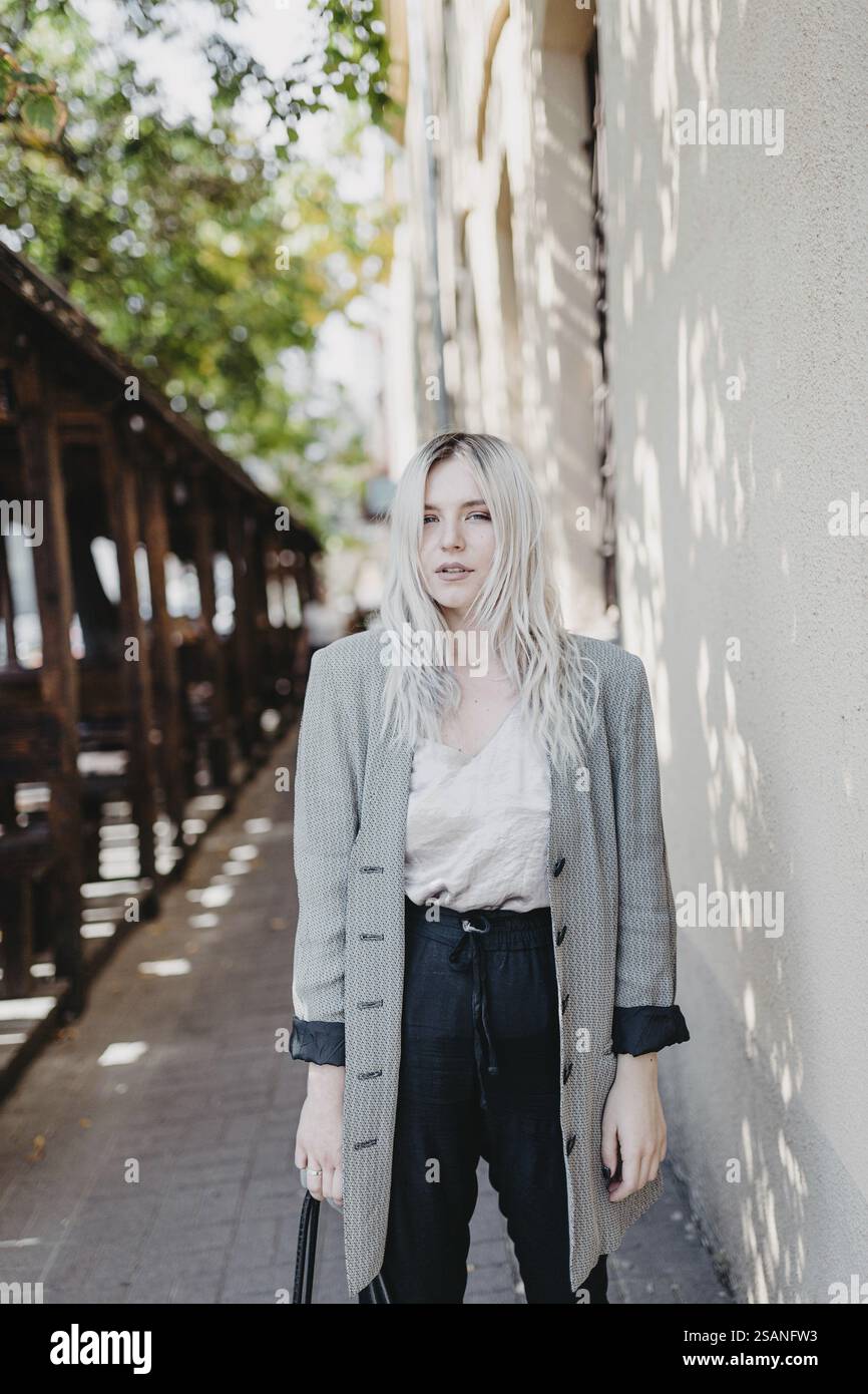 Una donna si trova su un sentiero illuminato dal sole e indossa abiti casual e un blazer grigio Foto Stock