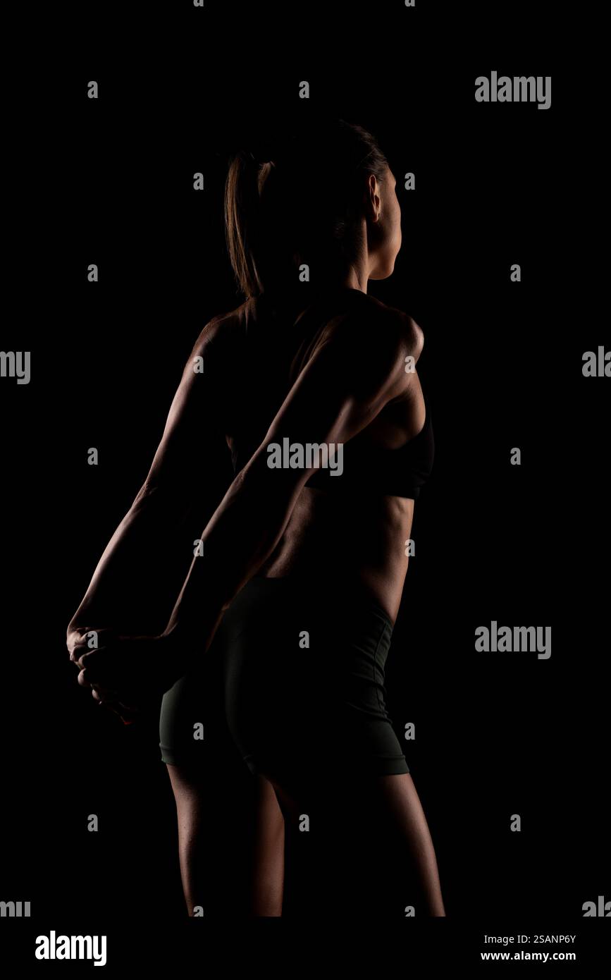 Silhouette di una donna impegnata in un esercizio di stretching, che enfatizza il tono muscolare e l'atletismo. Lo sfondo scuro evidenzia il movimento e cont Foto Stock