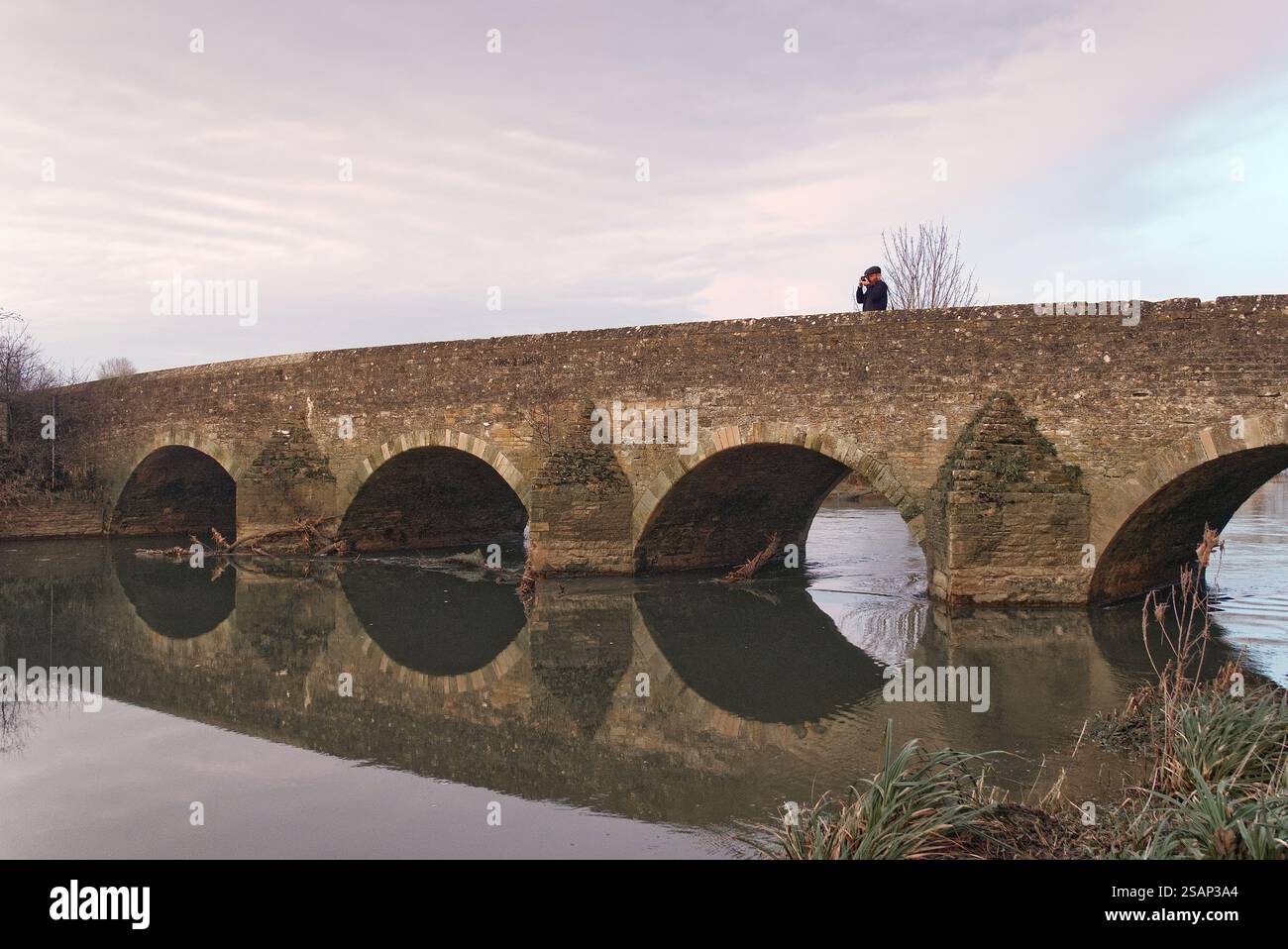 Uomo che scatta una foto dal ponte di Felmersham, Bedfordshire, Regno Unito, al tramonto, con riflessi nell'acqua del fiume Great Ouse Foto Stock