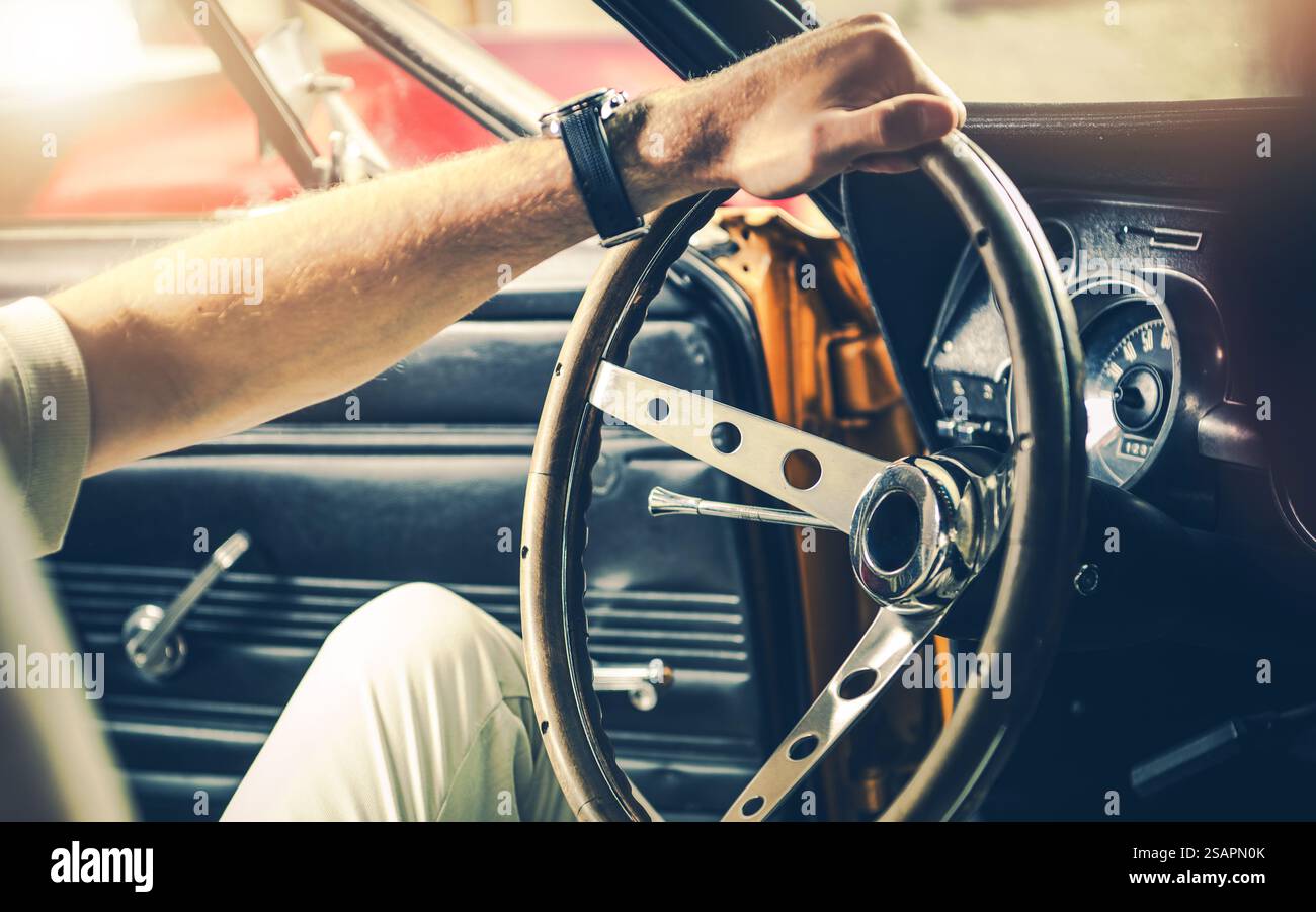 Un conducente tiene il volante d'epoca di un'auto d'epoca mentre si prepara a guidare attraverso un garage soffice al tramonto, mostrando il fascino del veicolo. Foto Stock