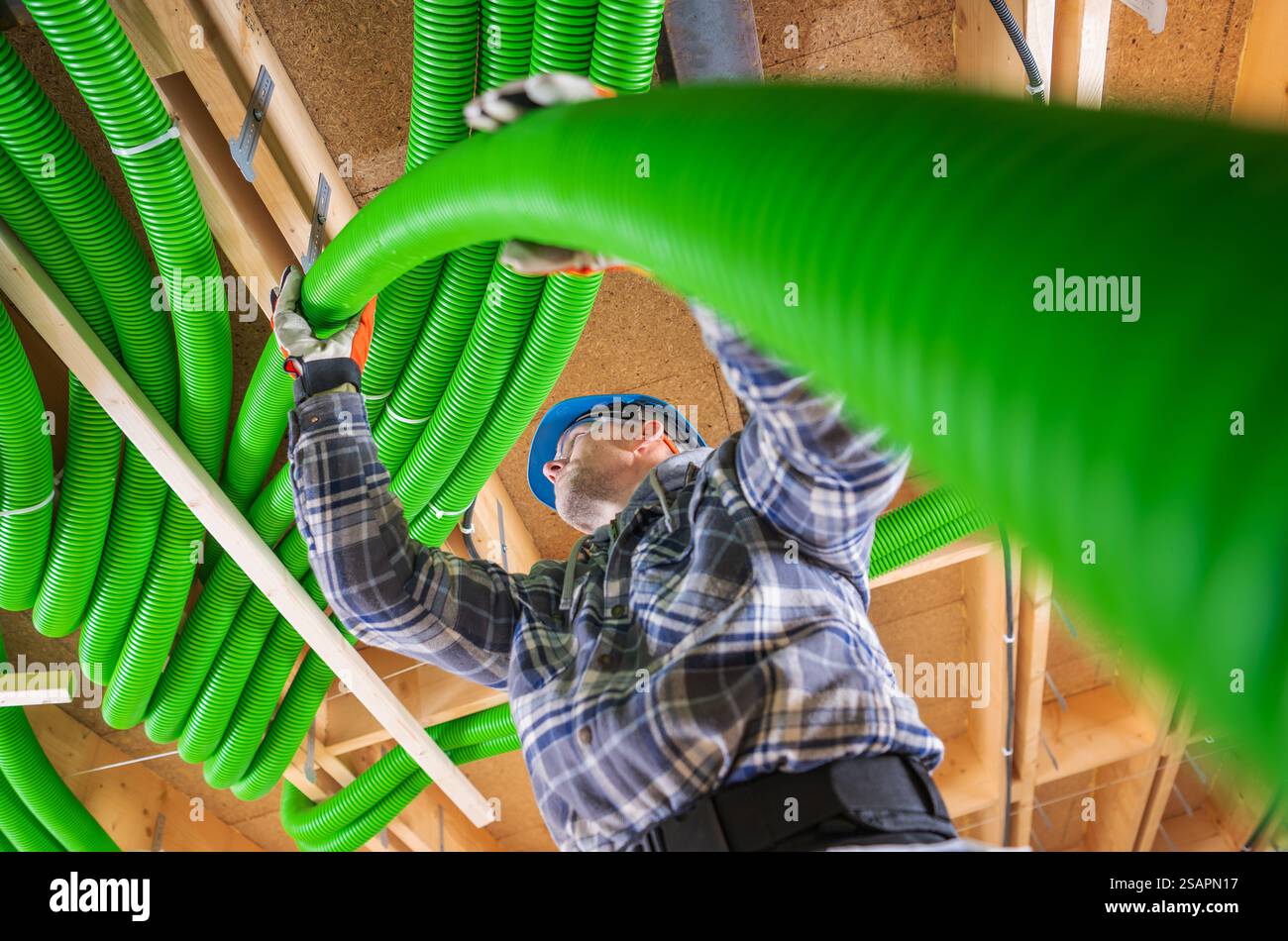 Un operaio edile sta installando un condotto verde sopra, indossando un casco e guanti in uno spazio interno ben illuminato. Concentrarsi sul lavoro di squadra e sul lavoro di squadra. Foto Stock