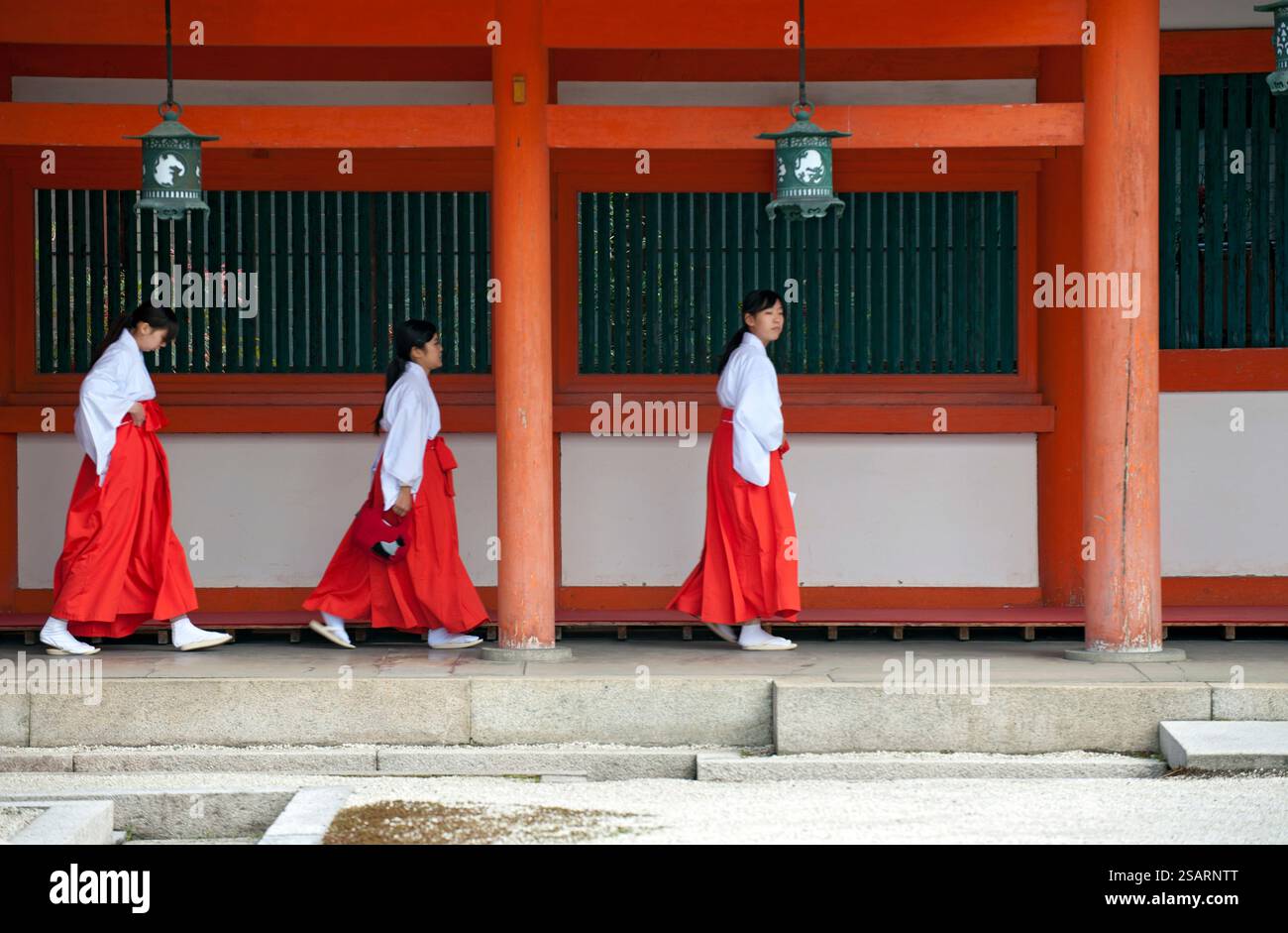 La fanciulla del Santuario ('miko') vestita con tradizionale hakama rosso e kosode bianco che cammina sotto un corridoio coperto al santuario shintoista Heian Jingu, Kyoto, Giappone. Foto Stock