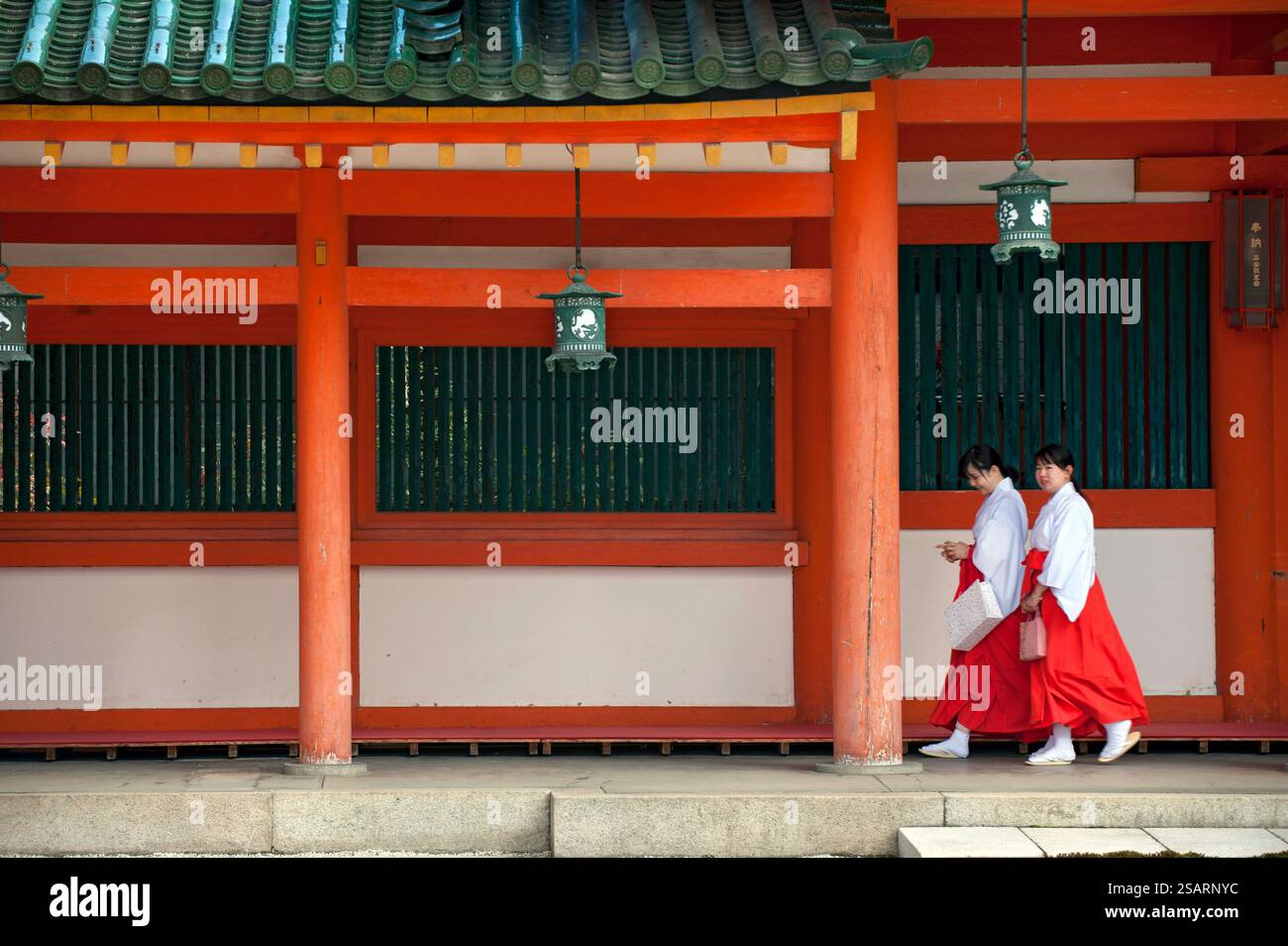 La fanciulla del Santuario ('miko') vestita con tradizionale hakama rosso e kosode bianco che cammina sotto un corridoio coperto al santuario shintoista Heian Jingu, Kyoto, Giappone. Foto Stock