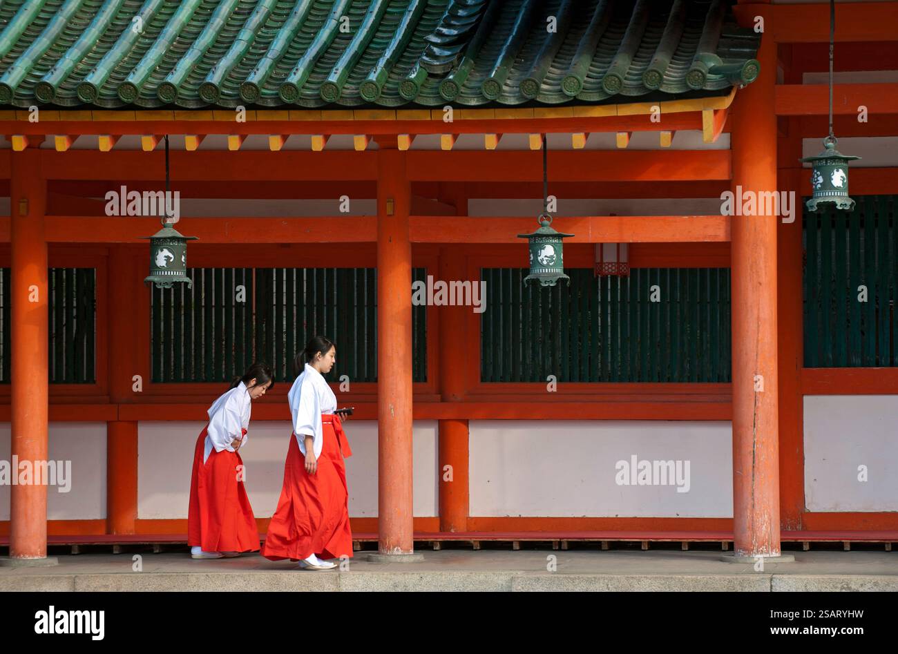 La fanciulla del Santuario ('miko') vestita con tradizionale hakama rosso e kosode bianco che cammina sotto un corridoio coperto al santuario shintoista Heian Jingu, Kyoto, Giappone. Foto Stock