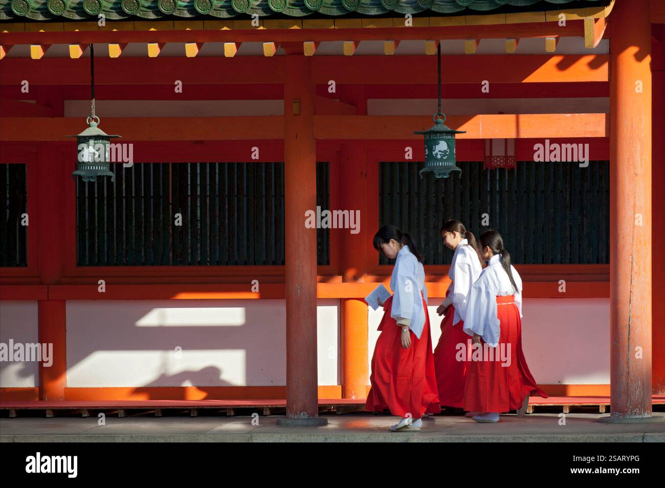 La fanciulla del Santuario ('miko') vestita con tradizionale hakama rosso e kosode bianco che cammina sotto un corridoio coperto al santuario shintoista Heian Jingu, Kyoto, Giappone. Foto Stock