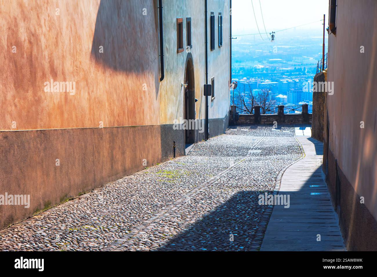 Stretto vicolo acciottolato fiancheggiato da alti edifici con vista su un paesaggio urbano lontano sullo sfondo. Il vicolo è parzialmente ombreggiato, con un tappeto di pietra Foto Stock