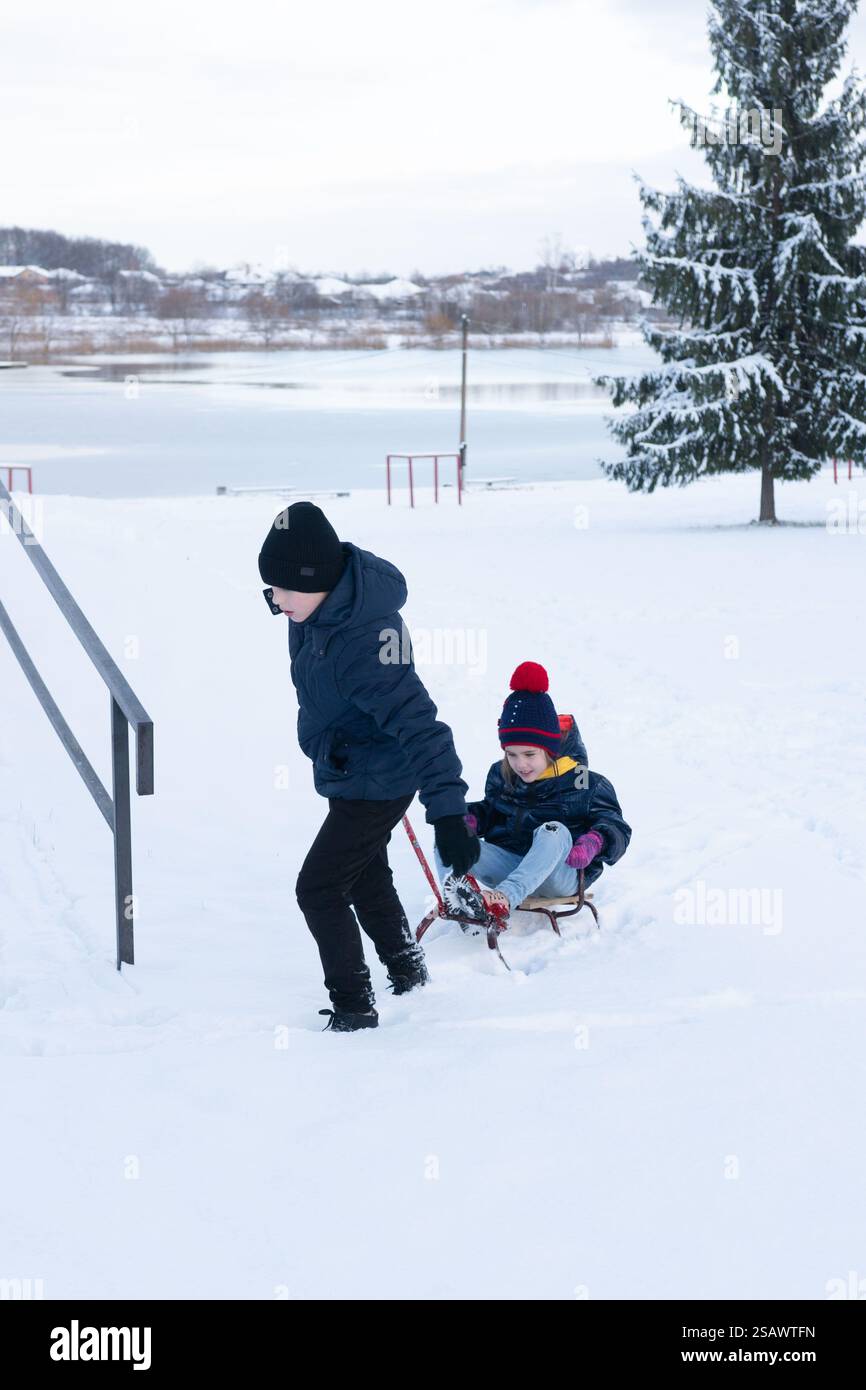 Due bambini trascorrono una giornata innevata. Un bambino tira una slitta con un altro bambino seduto su di essa, vicino a un lago parzialmente ghiacciato e a alberi innevati. La scena Foto Stock