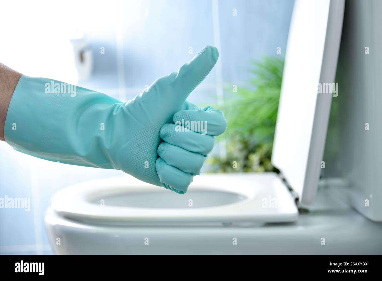 Bagno appena disinfettato con mano con guanti in gomma blu che danno il pollice verso l'alto gesto e ciotola WC sullo sfondo con coperchio aperto blu in piastrelle b Foto Stock