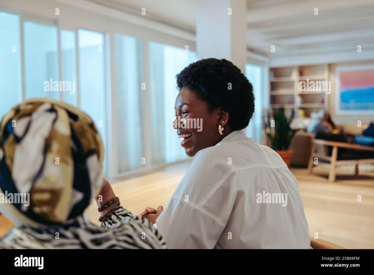 Una donna d'affari africana condivide una conversazione gioiosa con un collega in un ufficio aperto. Il suo comportamento amichevole e l'ambiente contemporaneo sottolineano Foto Stock