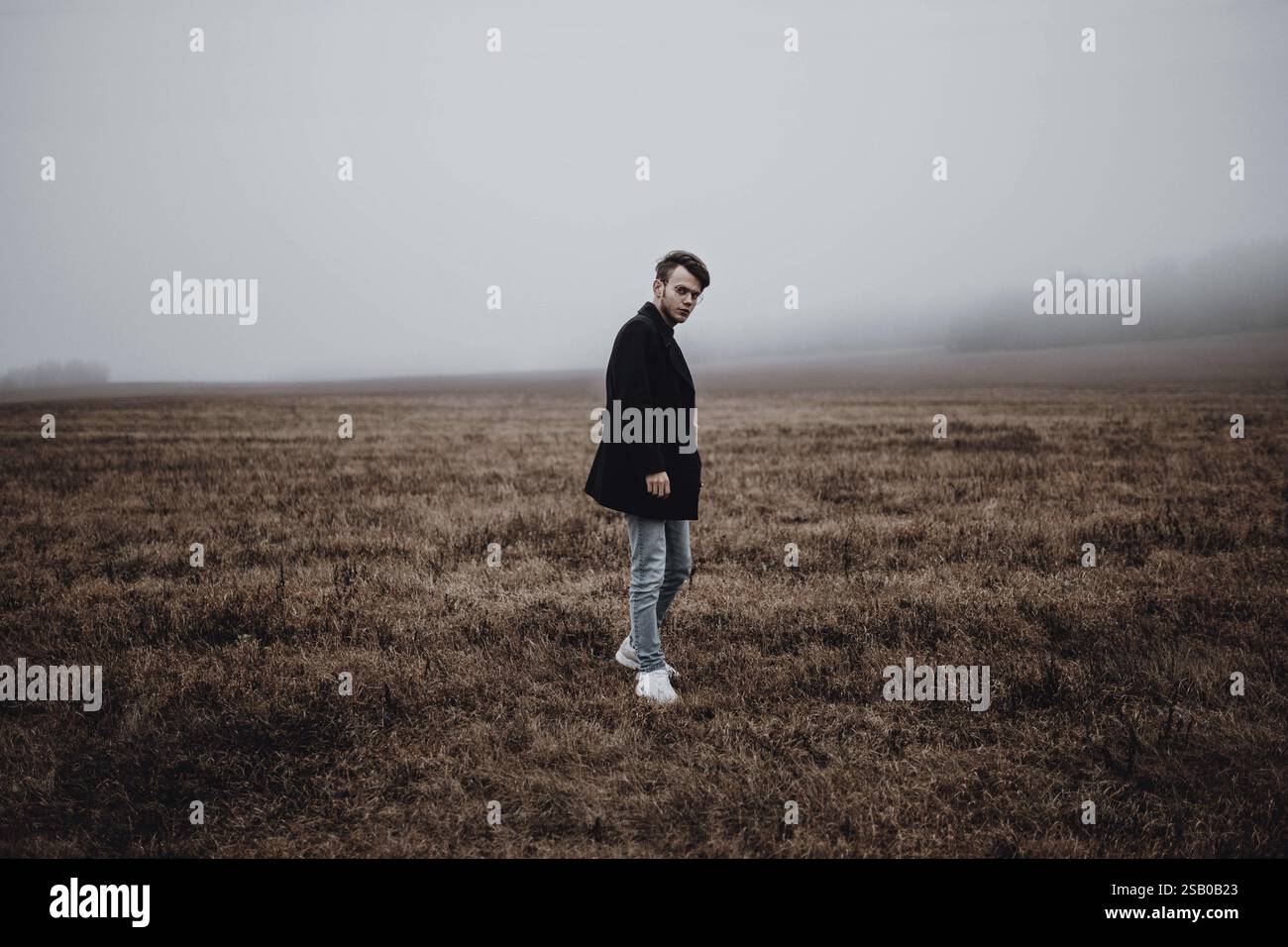 Un uomo contemplativo si trova in un campo nebbioso e indossa un cappotto, creando un'atmosfera cupa Foto Stock