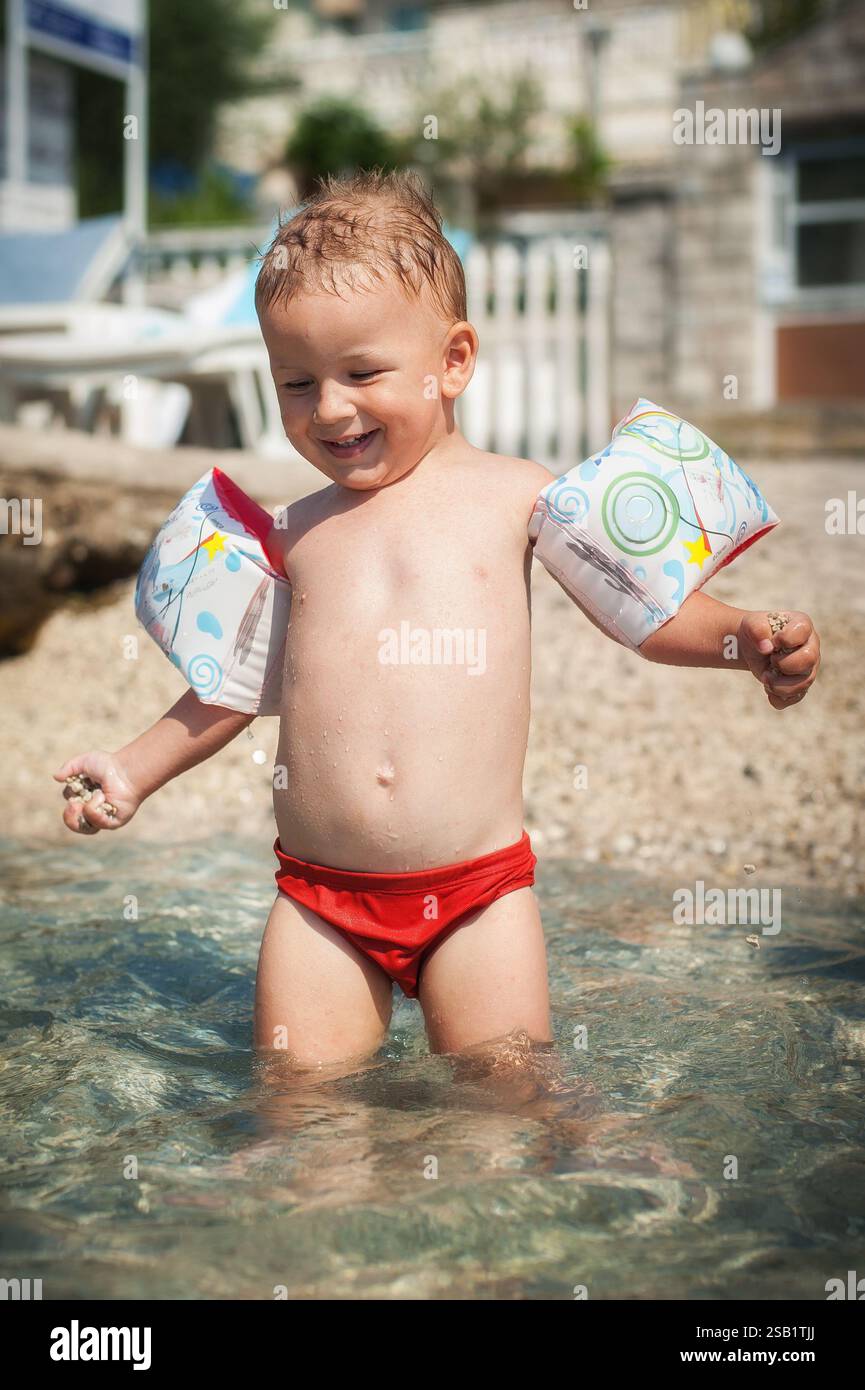 Un bambino allegro con i galleggianti gonfiabili per le braccia sta spruzzando in acque poco profonde sulla spiaggia. Il sole splende brillantemente, creando un'atmosfera allegra per su Foto Stock