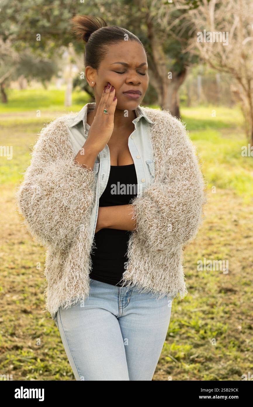Una donna nera con un abito casual si trova all'aperto, tenendo la guancia con un'espressione dolorosa, suggerendo disagio o mal di denti. Foto Stock