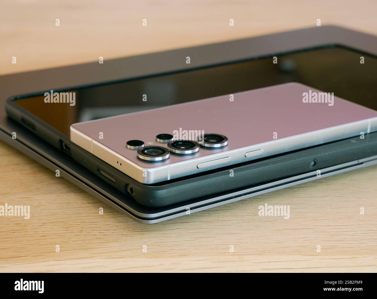 Un nuovo telefono cellulare argentato si trova sopra un tablet nero e un tablet si trova su un notebook argentato. Il portatile è su un tavolo di legno. Foto Stock