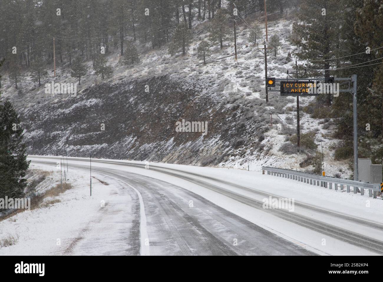 Una tempesta invernale copre la State Route 36 nella contea di Lassen, California, con neve e ghiaccio, poiché un segnale stradale avvisa i conducenti delle curve ghiacciate che precedono per sicurezza. Foto Stock