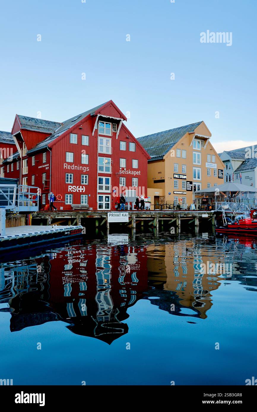 Gli edifici del porto di Tromso, Norvegia, riflettono sulle acque calme al tramonto, mostrando un'architettura colorata e una barca di salvataggio attraccata Foto Stock