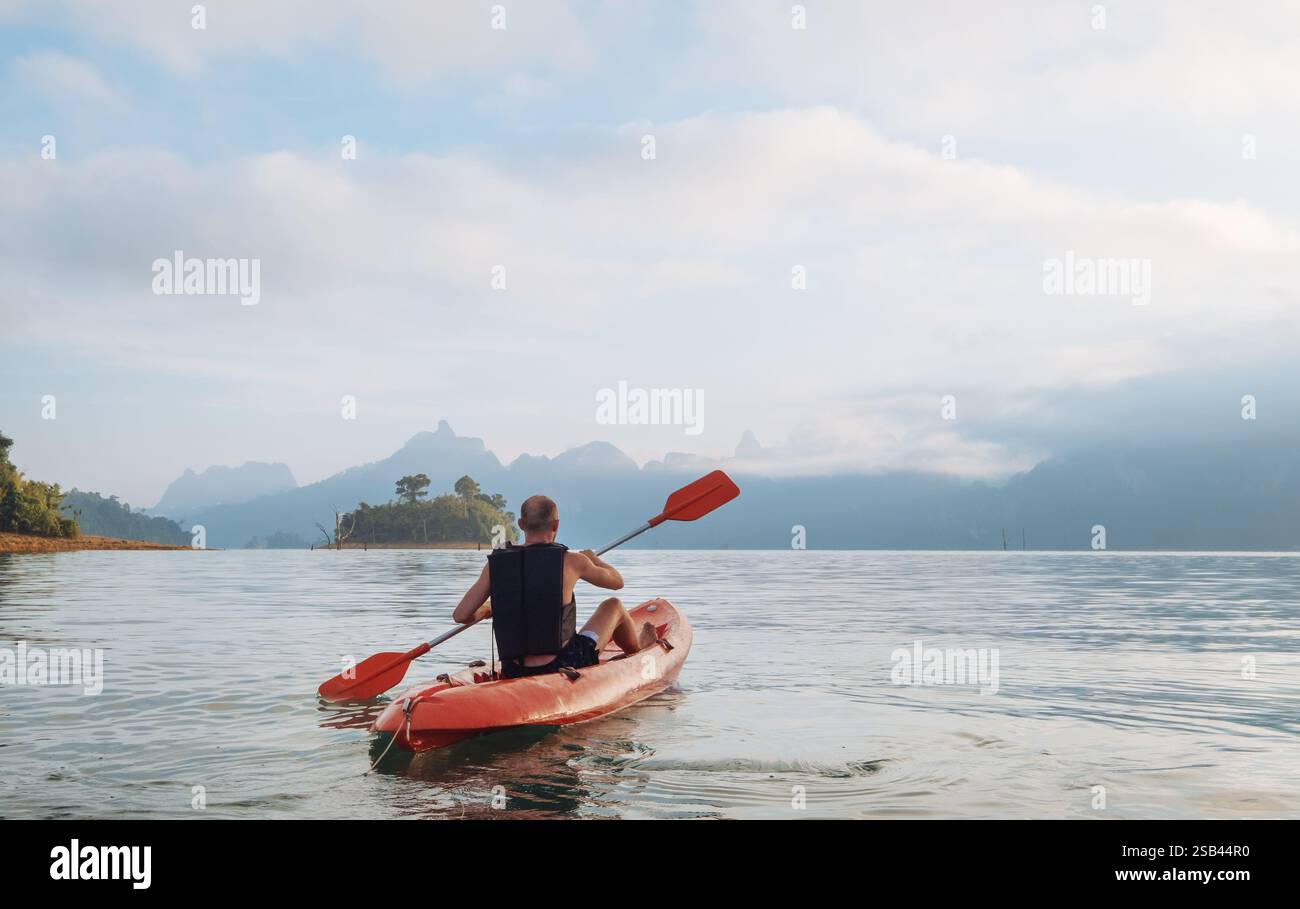 Uomo che pagaia in kayak sul calmo lago Cheow LAN e incontra l'alba nel parco nazionale di Khao Sok, Thailandia. Acque tranquille, luce dorata e scogliere calcaree creano Foto Stock