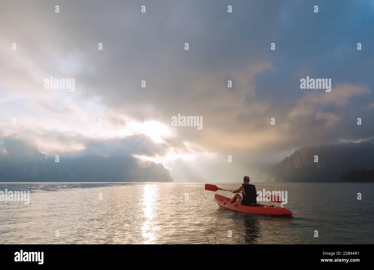 Uomo che pagaia in kayak sul calmo lago Cheow LAN e incontra l'alba nel parco nazionale di Khao Sok, Thailandia. Acque tranquille, luce dorata e scogliere calcaree creano Foto Stock