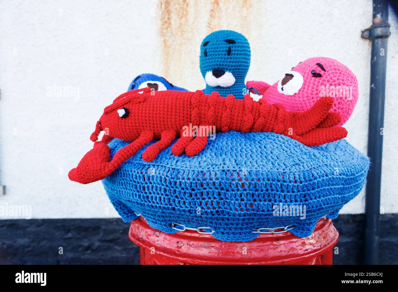 Colorate creature marine di lana all'uncinetto, polpo di aragosta esposto su un vibrante cuscino in maglia su una cassetta per posta rossa contro una parete testurizzata Foto Stock