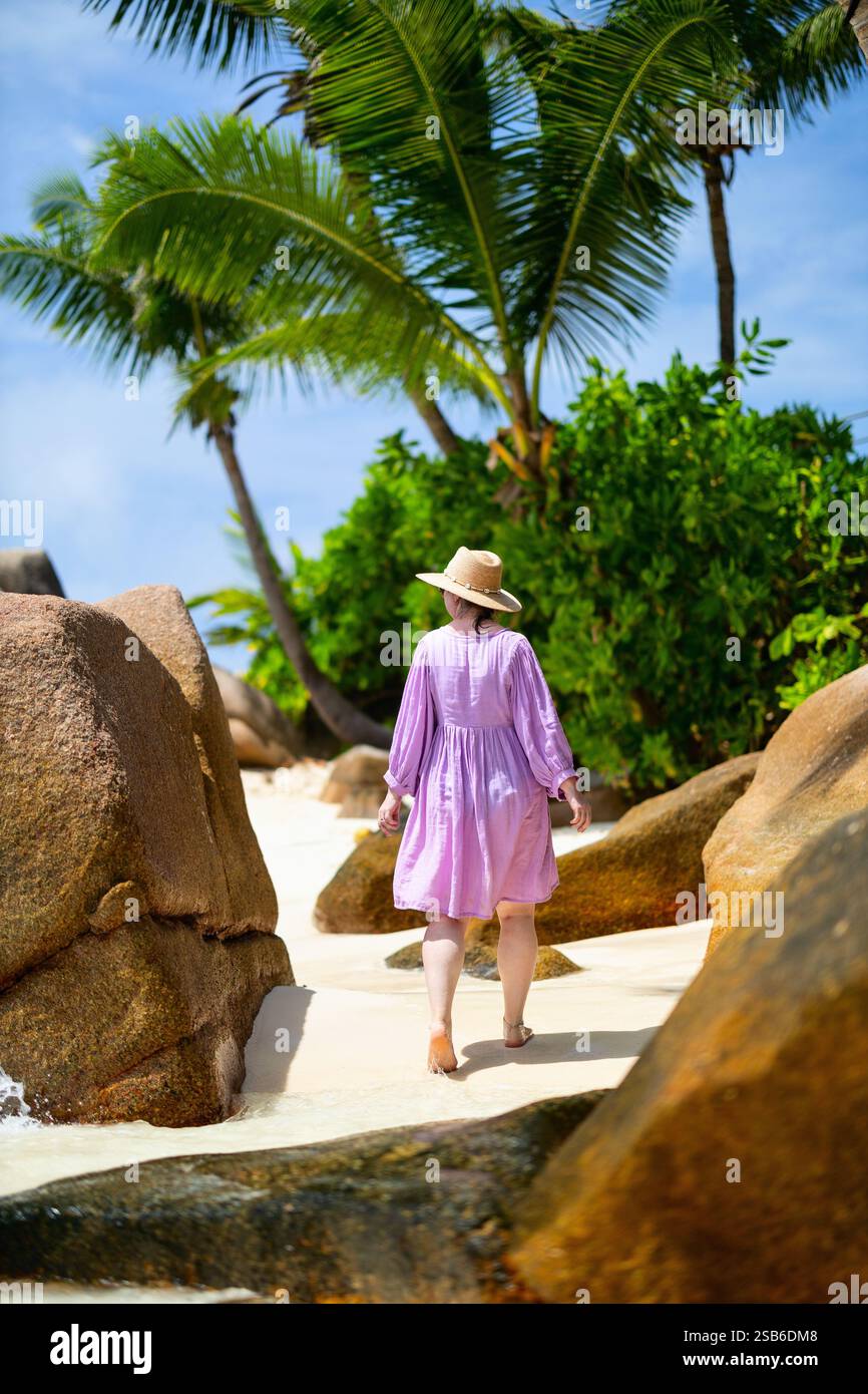 Una donna in abito rosa e cappello da sole che cammina a piedi nudi su una spiaggia tropicale, circondata da grandi rocce, palme e vegetazione lussureggiante Foto Stock