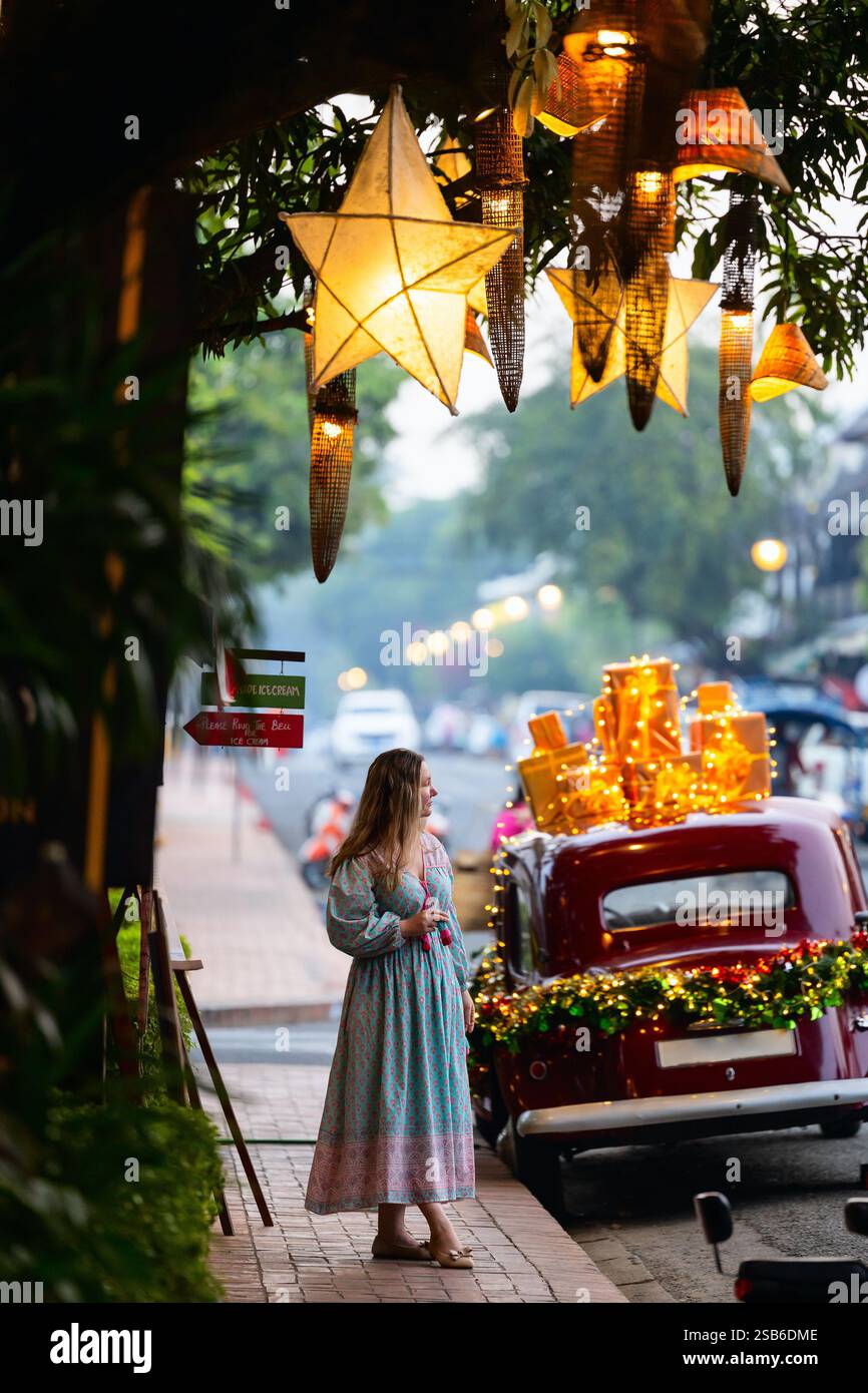 Una donna si erge su una strada decorata con lanterne scintillanti delle stelle natalizie appese sopra e un'auto d'epoca adornata con scatole regalo e luci festive Foto Stock Una donna si erge su una strada decorata con lanterne scintillanti delle stelle natalizie appese sopra e un'auto d'epoca adornata con scatole regalo e luci festive Foto Stock