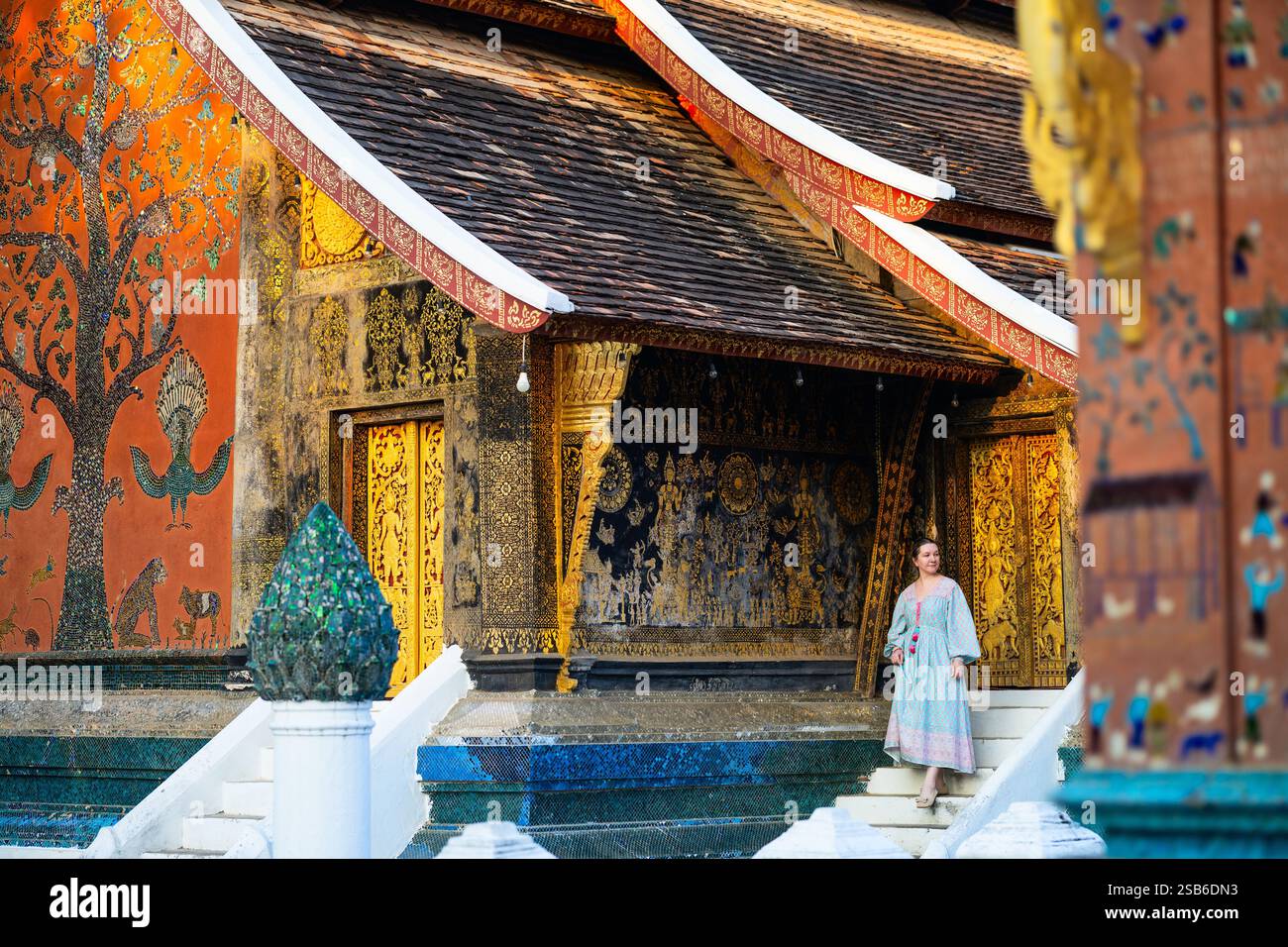 Una donna cammina vicino alle pareti ornate di Wat Xiengthong a Luang Prabang, Laos, caratterizzato da un dettagliato murale di alberi a mosaico e intagli dorati Foto Stock