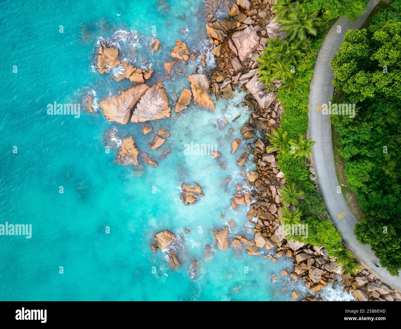 Vista aerea della costa dell'isola di la Digue, Seychelles, che mostra acque turchesi, grandi massi sparsi, una strada tortuosa costeggiata da verde lussureggiante Foto Stock