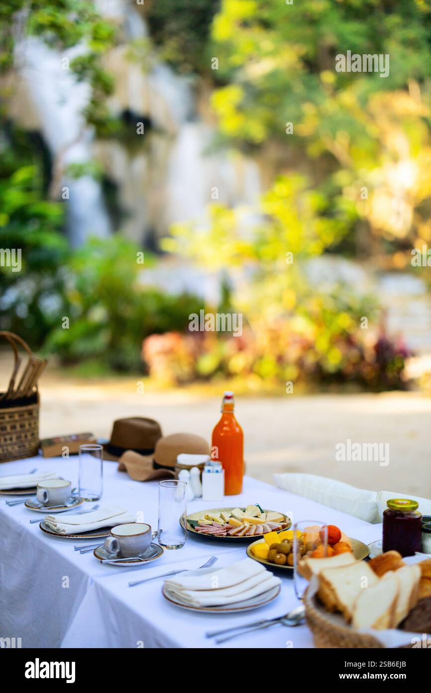 Un tavolo all'aperto e' allestito con prodotti per la colazione, che includono pane, frutta e succo di frutta, vicino ad una cascata panoramica circondata da vegetazione lussureggiante Foto Stock