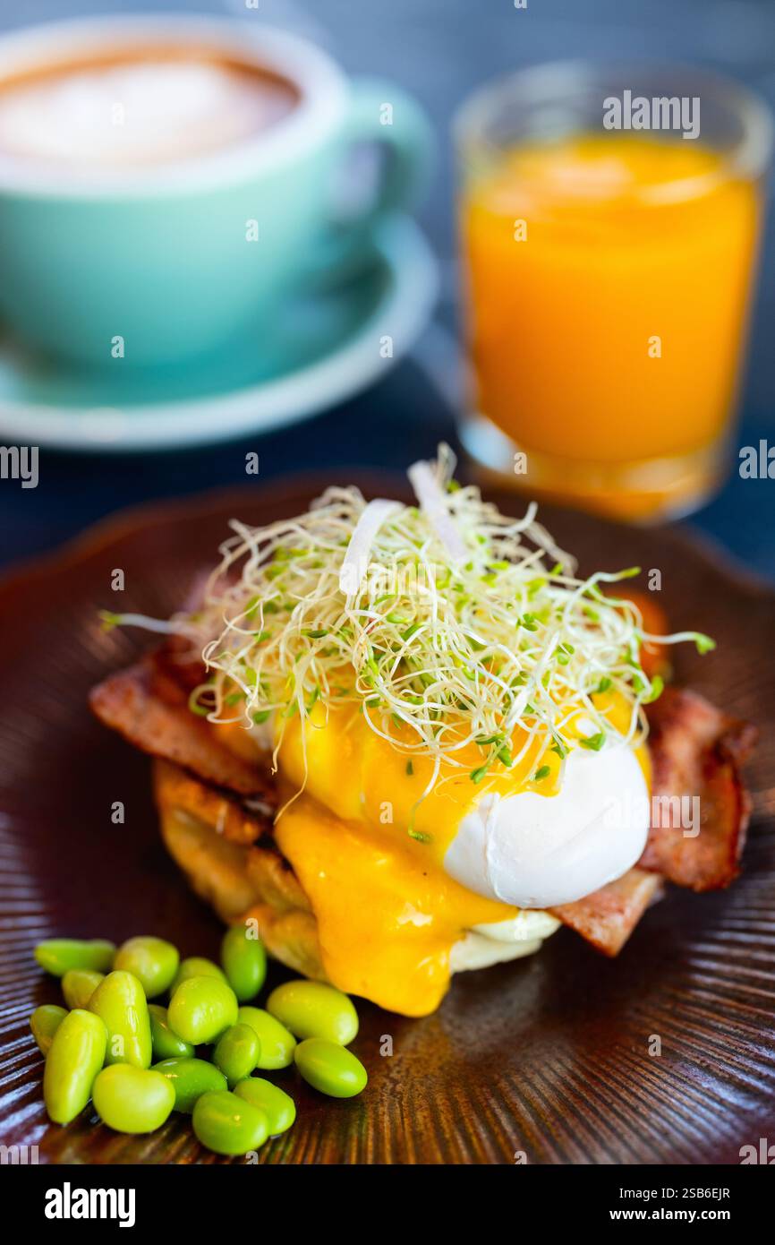 Una porzione gourmet di uova Benedict condite con germogli freschi e cremosa salsa hollandaise, accompagnata da edamame, con cappuccino e succo d'arancia Foto Stock