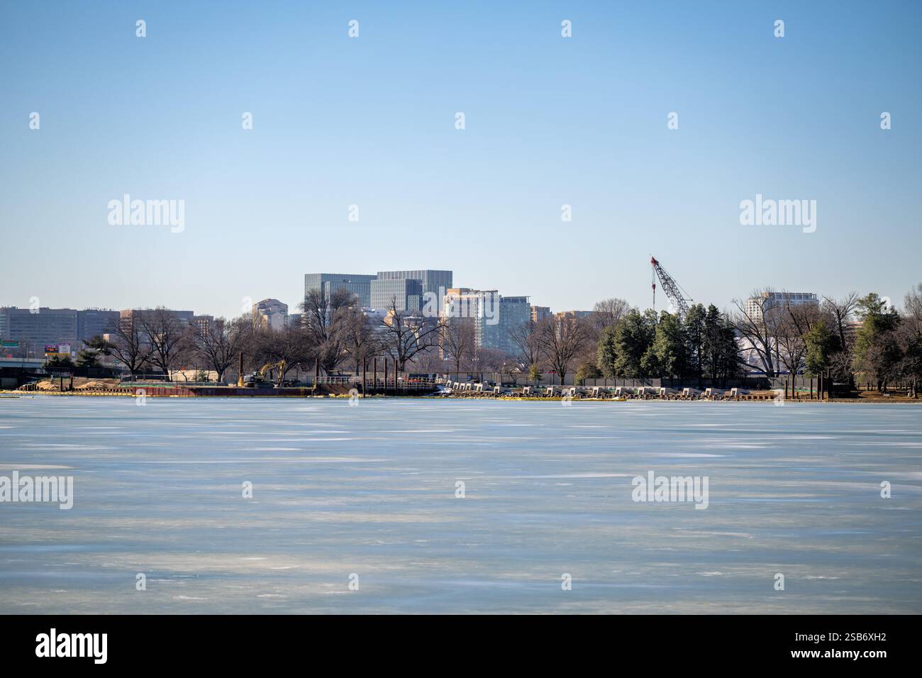 WASHINGTON DC - Un bacino di marea ghiacciato è visto in un giorno d'inverno, con lo skyline di Rosslyn, Virginia, visibile sullo sfondo. Gli alberi dormienti lungo la riva si stagliano in contrasto con la loro apparizione durante l'annuale National Cherry Blossom Festival in primavera. Le attrezzature da costruzione sono visibili lungo la banchina. Foto Stock