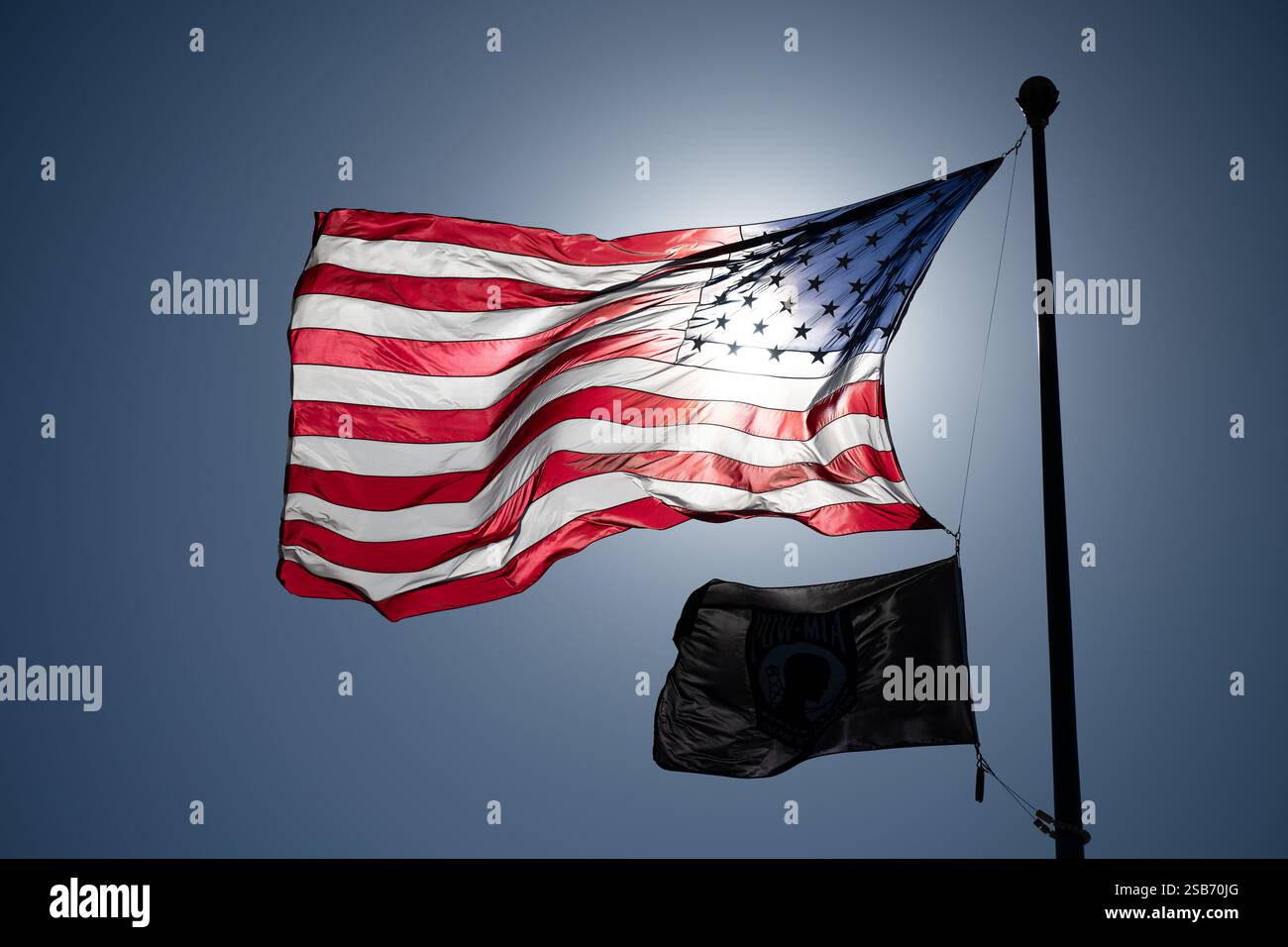 WASHINGTON DC - una bandiera americana e una bandiera POW/mia volano insieme contro un cielo azzurro, retroilluminato dal sole a Washington DC. La bandiera nera POW/mia, posizionata sotto le stelle e le strisce, commemora i prigionieri di guerra e i dispersi in azione. Questa mostra patriottica è una vista comune degli edifici federali e dei memoriali in tutta la capitale della nazione. Foto Stock