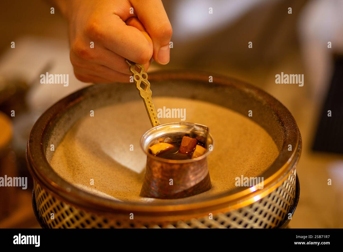La mano di un barista crea sapientemente il caffè sulla sabbia utilizzando una tradizionale cezve turca di rame, mostrando un metodo di estrazione unico in un caldo e rustico setti Foto Stock
