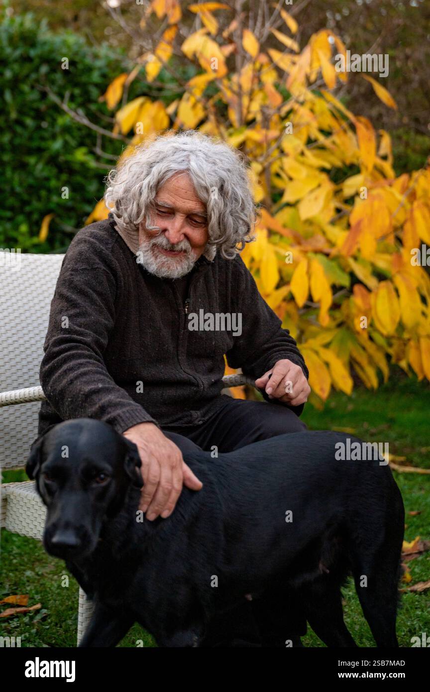 Un uomo anziano con i capelli ricci grigi e la barba si siede in un giardino, accarezzando delicatamente un Labrador nero. L'autunno dorato lascia alle spalle un caldo, Foto Stock