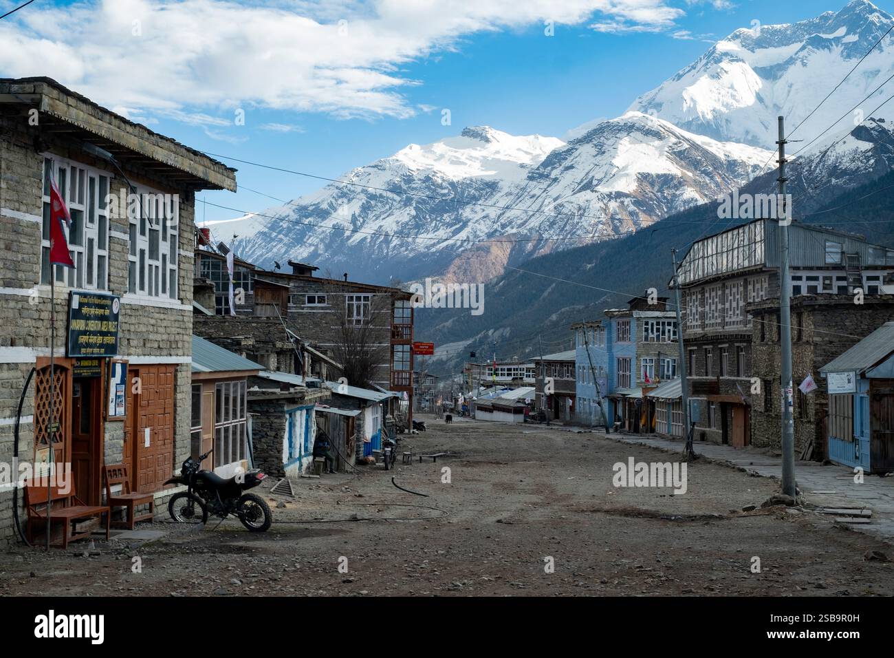 La strada principale di Manang, fiancheggiata da edifici in pietra e adagiata sullo sfondo di montagne innevate. Foto Stock