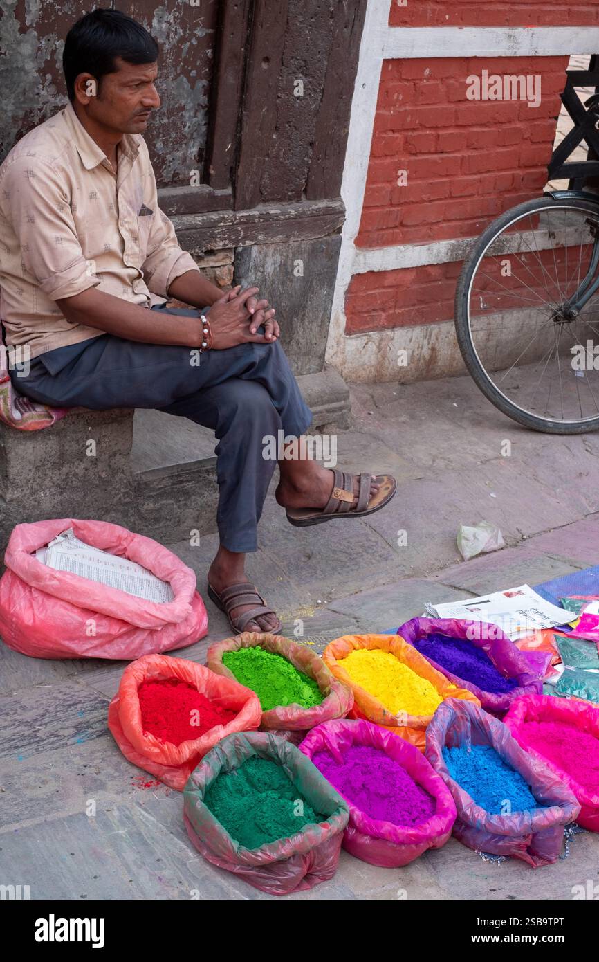 In mezzo al caos vibrante di Holi, un venditore con una camicia marrone offre polveri colorate, che riflettono lo spirito del festival con un comportamento tranquillo. Foto Stock