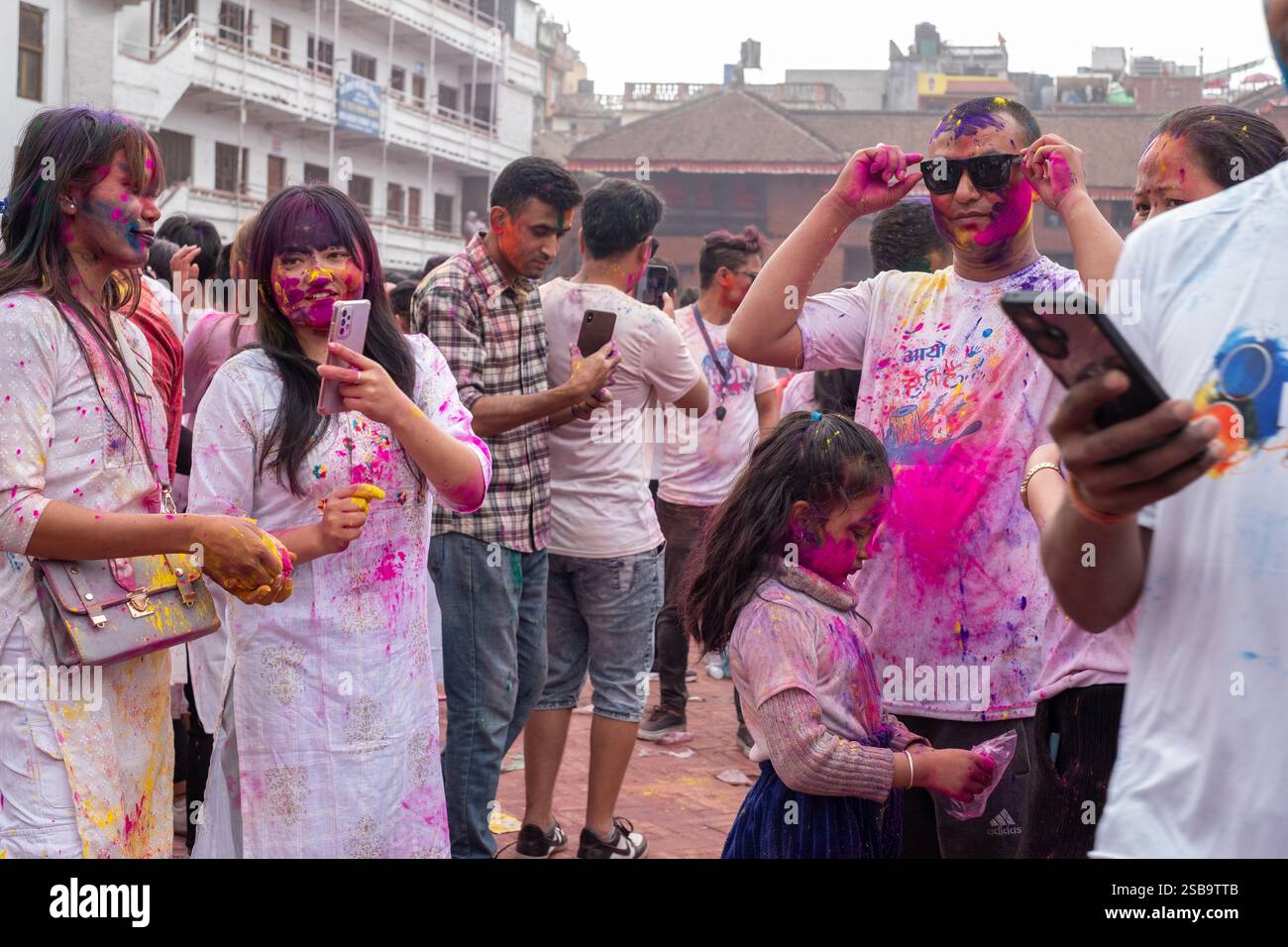 Gruppo di giovani nepalesi vestiti con abiti luminosi ricoperti di colori Holi, in piedi e chiacchierando con i loro cellulari a Durbar Square, Kathmandu, Nepal. Foto Stock