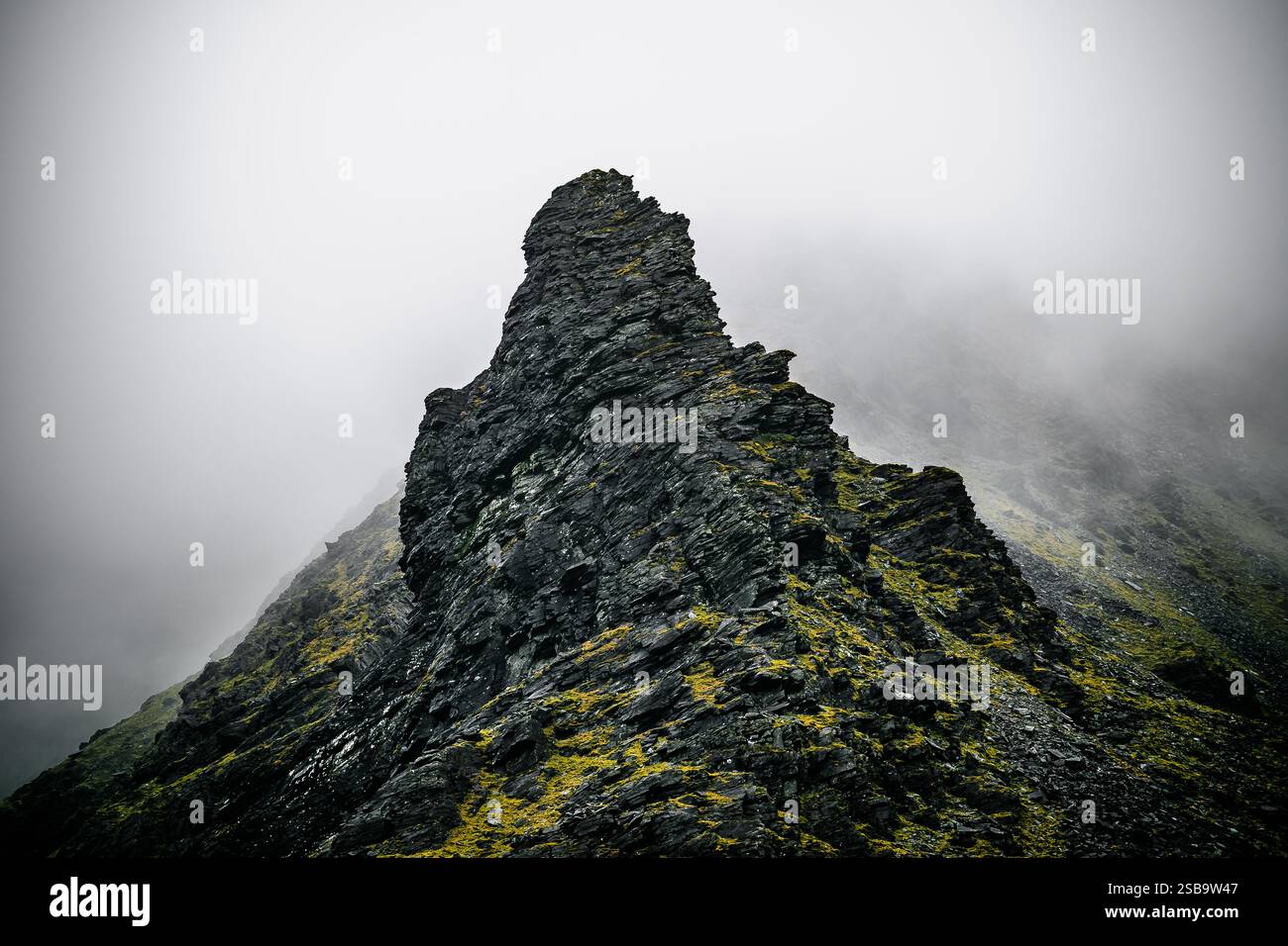 Un'aspra vetta rocciosa si innalza bruscamente in un cielo nuvoloso, circondato dalla nebbia. Le formazioni frastagliate sono adornate da macchie di muschio verde, che esaltano la W Foto Stock