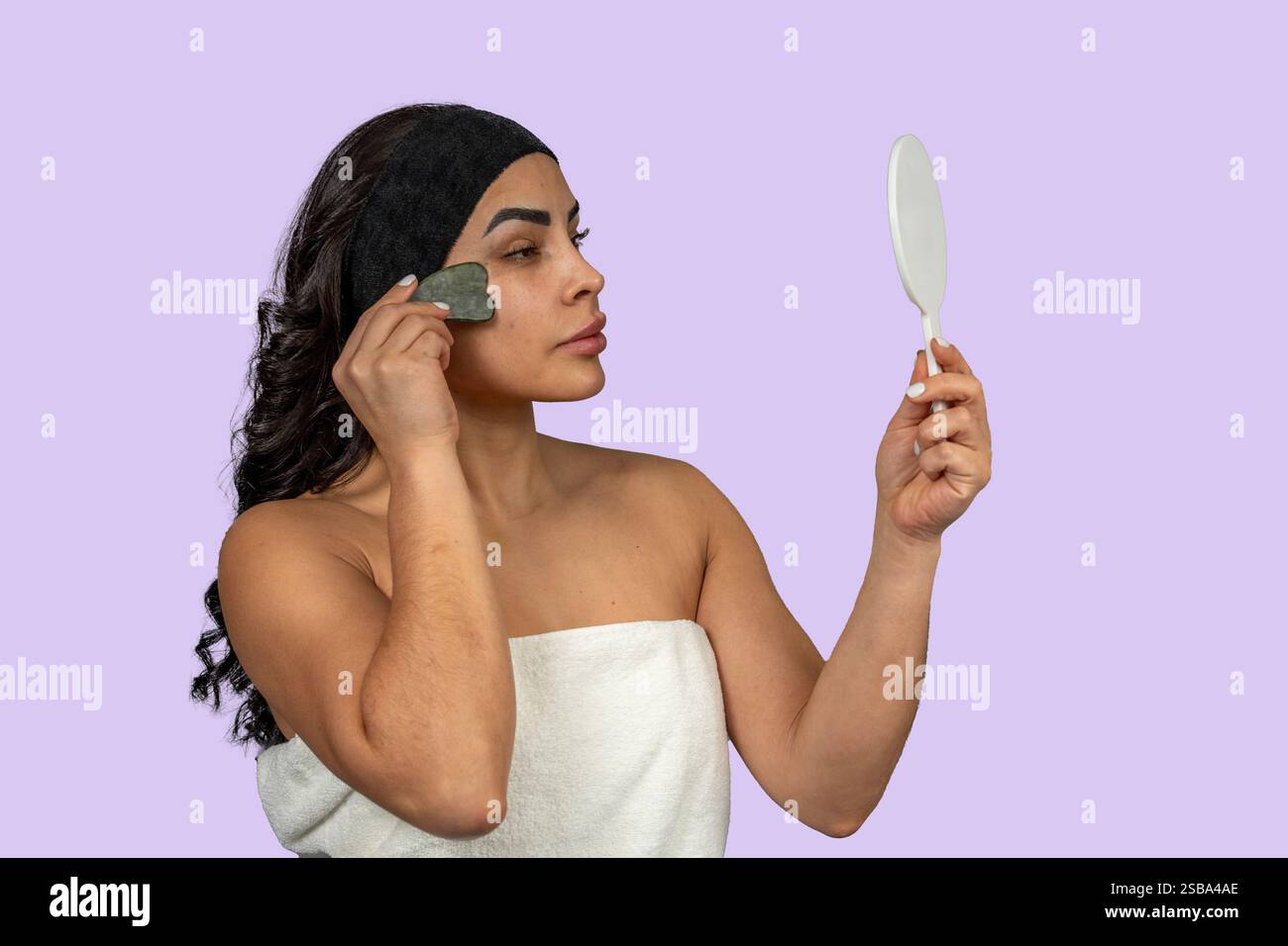 Donna che usa uno strumento di massaggio gua Sha sul viso e controlla i risultati con uno specchio per le mani Foto Stock