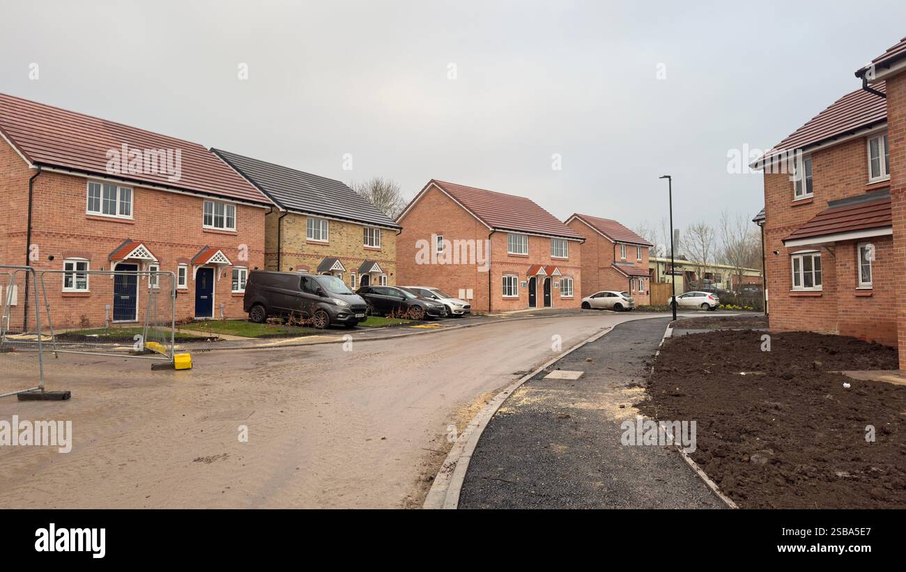 Case in mattoni di nuova costruzione fiancheggiano una strada fangosa, alcune con auto parcheggiate. Gli elementi di costruzione, compresa una recinzione temporanea, sono ancora visibili. Il Foto Stock