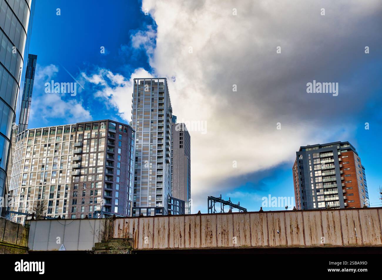 I moderni edifici di appartamenti si innalzano contro un cielo blu parzialmente nuvoloso. Una struttura arrugginita è in primo piano. Gli edifici variano in altezza, colore, Foto Stock