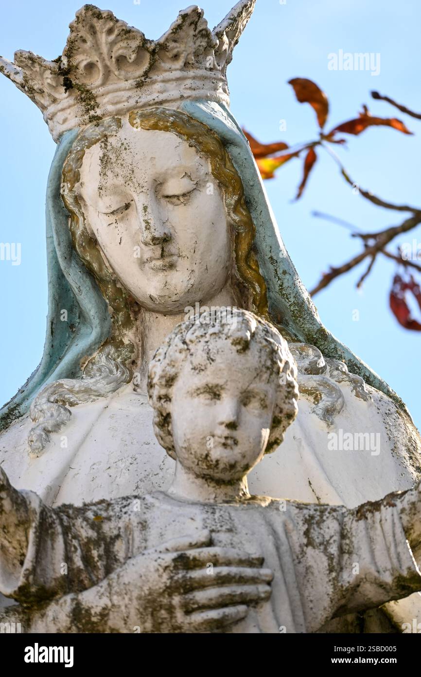 Primo piano di una statua della Madonna intemprata con una corona e un velo, che tiene in mano il bambino Gesù, incorniciata da un cielo blu e foglie d'autunno. Foto Stock
