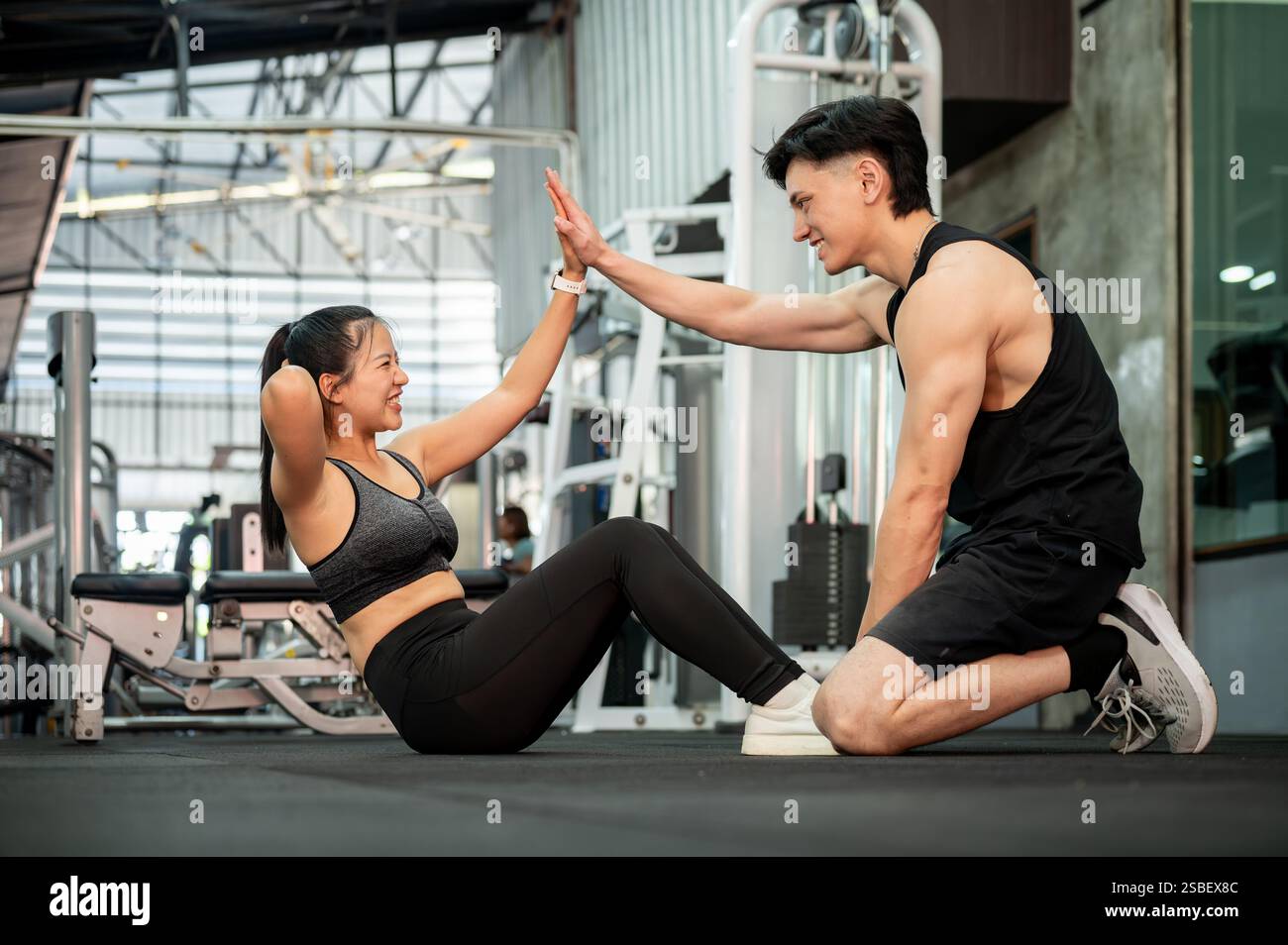Un simpatico e allegro allenatore di fitness maschile asiatico sta dando un alto cinque mentre istruisce la sua cliente femminile su una forma di sit-up adeguata in palestra. healt Foto Stock