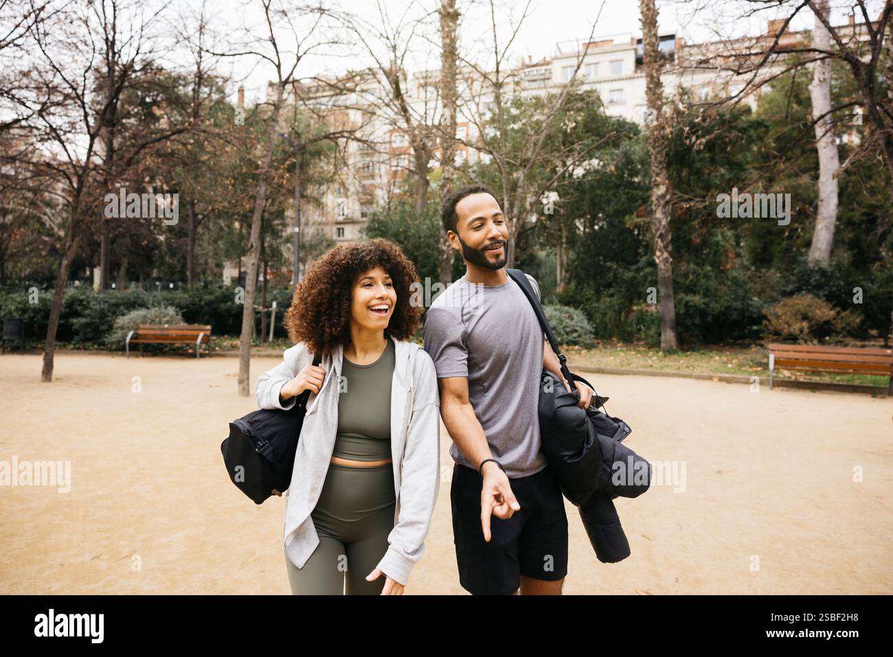Il personal trainer e il suo tirocinante camminano all'aperto in un parco dopo una sessione di allenamento in un parco urbano, trasportando borse e indossando abiti da fitness. Foto Stock