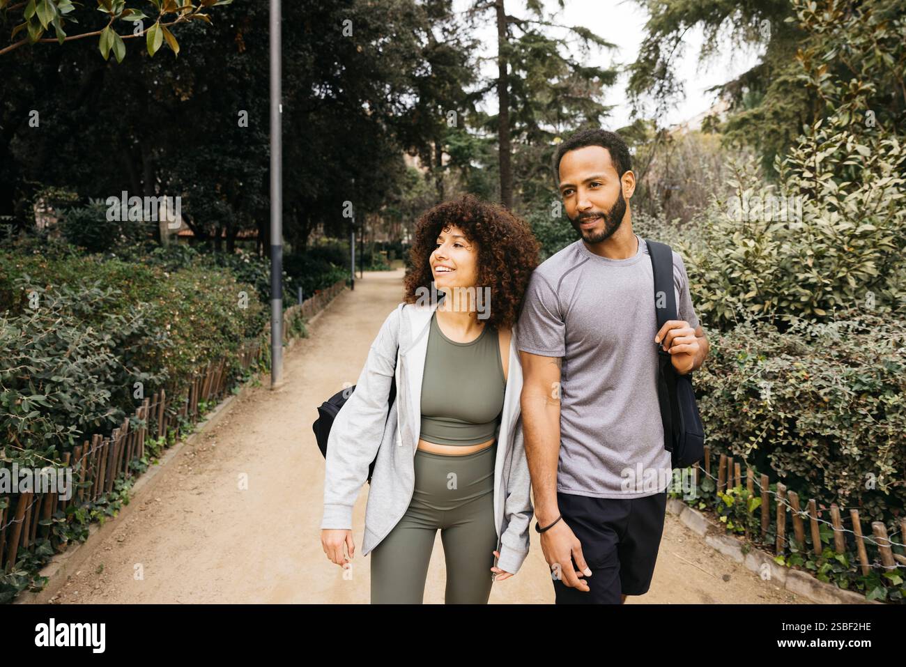 Una coppia allegra passeggia lungo un bellissimo sentiero del parco con alberi e piante lussureggianti, indossando abbigliamento fitness e portando con sé borse da palestra. Foto Stock