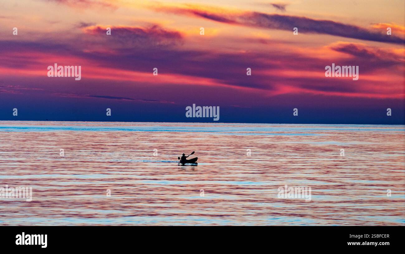 Un tramonto sereno sulle acque calme, caratterizzato da un kayak solitario con vivaci sfumature di viola, arancione e rosa nel cielo. Foto Stock