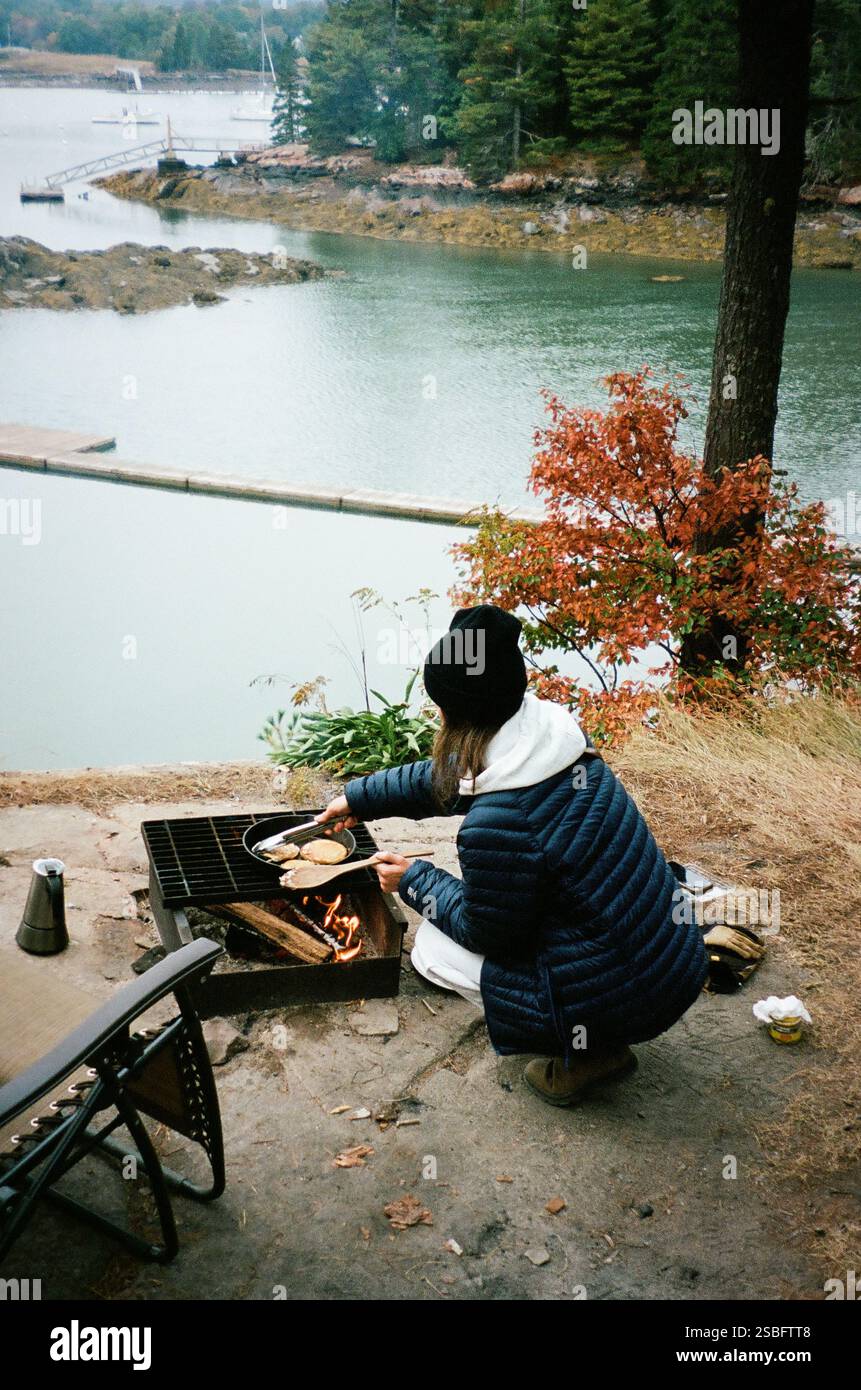 Donna che cucina sopra un fuoco da campo vicino a un lago panoramico in autunno. Foto Stock