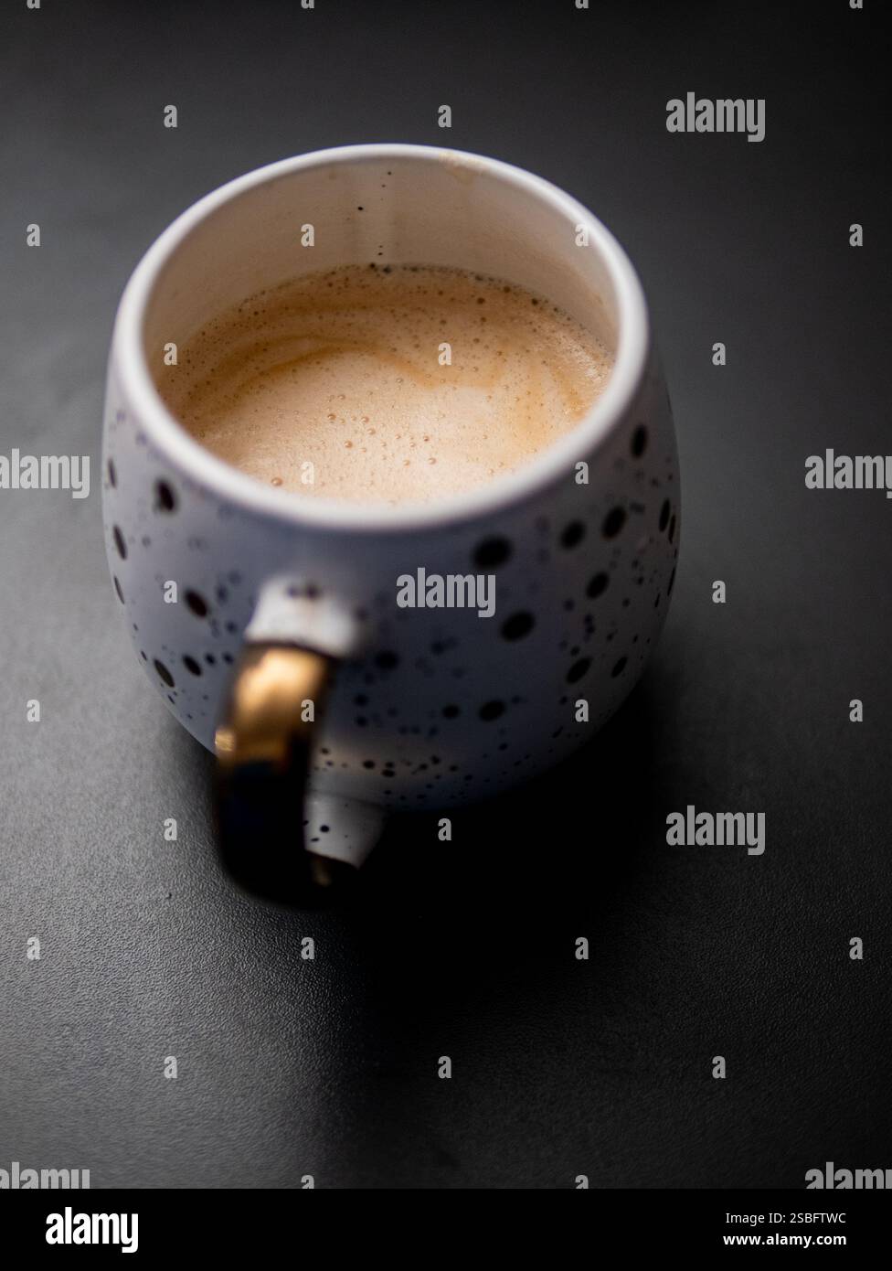 Una tazza da caffè bianca con manico dorato e macchie nere. Il caffè è marrone e schiumoso Foto Stock